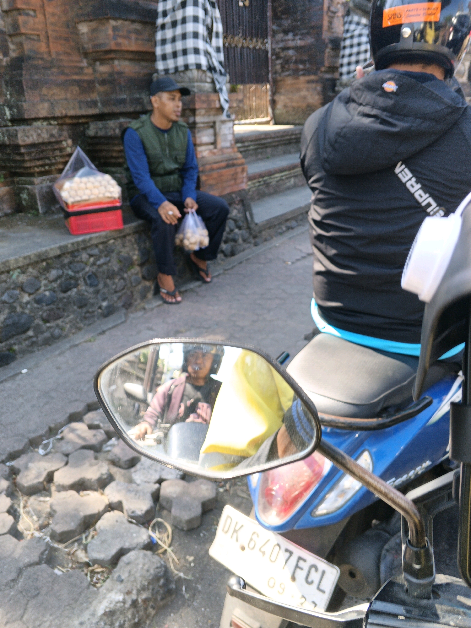 In the city of Abiansemal, Indonesia, a person is seated on a stone ledge next to a brick building. They are wearing a dark baseball cap, a green vest over a blue long-sleeved shirt, and dark pants. In front of them, a red container holds a large bag of snacks. The person is holding a plastic bag with snacks in their right hand. To the right, another person wearing a black jacket and a black helmet is seated on a blue motorcycle. The motorcycle's side mirror reflects a person wearing a dark jacket and a yellow garment, possibly the motorcycle rider. A license plate on the motorcycle reads "DK 6407FCL". The environment appears to be an outdoor area with paved ground made of interlocking concrete blocks.