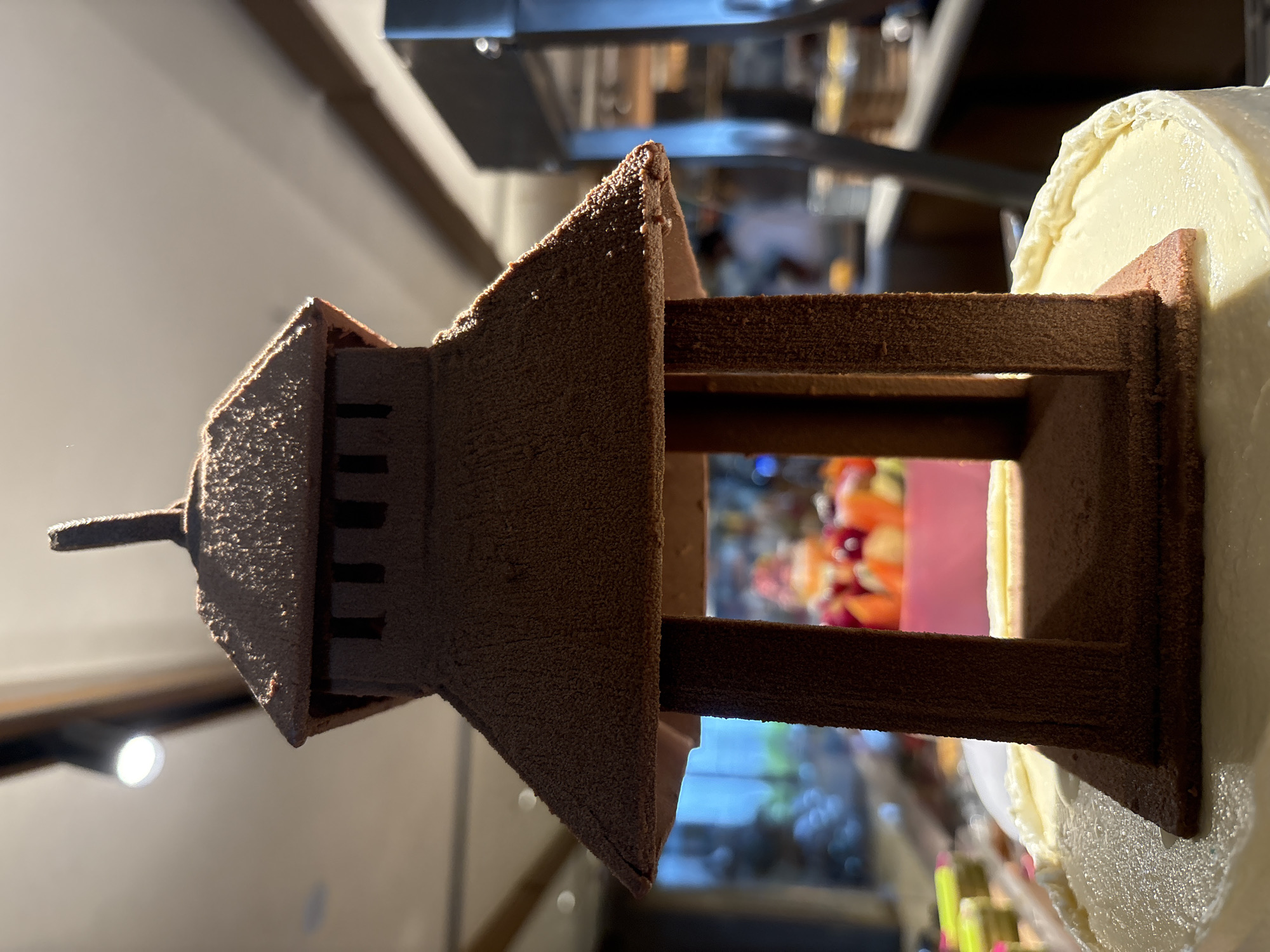 A close-up, low-angle shot features a chocolate lantern-like dessert sitting atop a white frosted cake. The dessert is dark brown and textured, resembling a miniature pagoda or traditional lantern with a pointed roof and rectangular openings. The cake is a pale yellow or cream color with a smooth, slightly textured frosting. In the blurred background, a display of colorful fruit can be seen, along with other food items and what appears to be a buffet setting. The lighting suggests an indoor environment, possibly a restaurant or banquet hall.