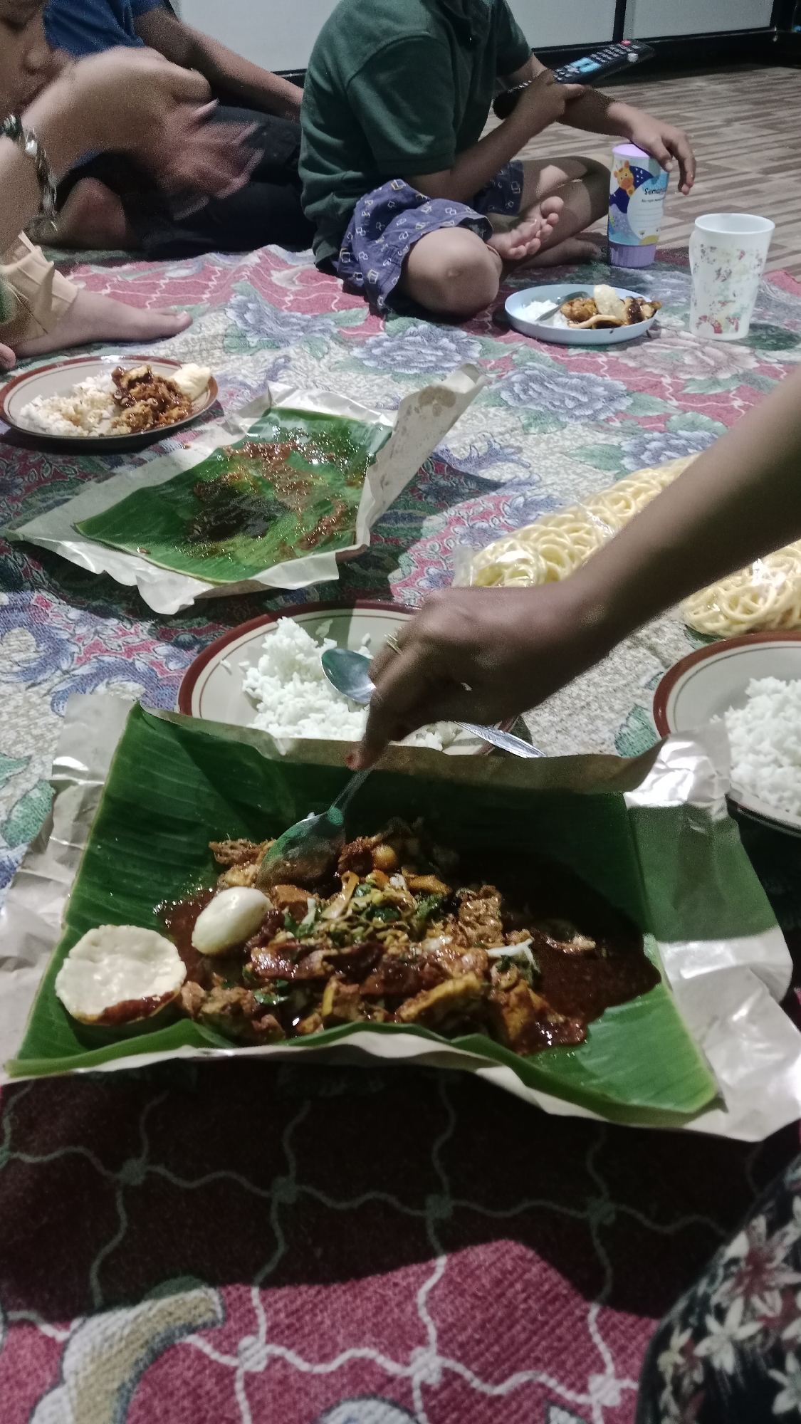 A group of people are sitting on the floor, eating from plates of food laid out on a patterned mat. The food is served on banana leaves, with rice, meat, and vegetables visible. Some of the people are children, and one is holding a remote control. The scene appears to be a casual meal at a home in Madiun, Indonesia, given the traditional serving style of food on banana leaves.