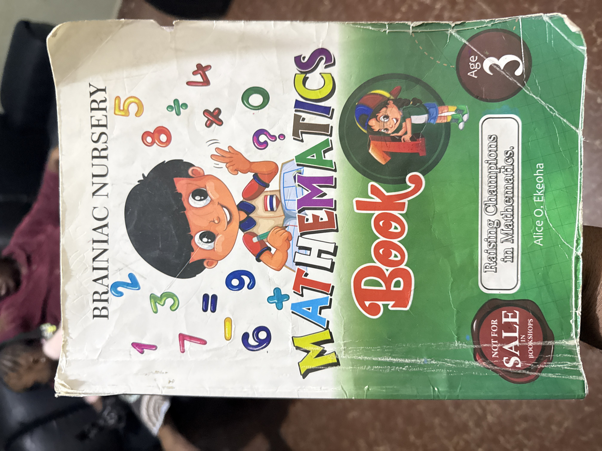 A slightly worn children's mathematics textbook, titled "BRAINIAC NURSERY MATHEMATICS Book 1," is held vertically in the foreground. The upper half of the cover is white, featuring a cartoon boy drawing in a notebook, surrounded by colorful numbers (1 through 9, 0) and basic mathematical symbols (+, -, =, x, ÷, ?). The lower half is green with a subtle grid pattern. The word "MATHEMATICS" is prominently displayed in large, multi-colored block letters. Below this, "Book 1" is rendered in a red, bubbly font, with a cartoon girl holding the number "1" inside the '1' of 'Book 1'.
A circular red seal in the bottom left corner states "NOT FOR SALE IN BOOKSHOPS". A white rectangular box below the main title reads "Raising Champions in Mathematics.". The author's name, "Alice O. Ekeoha", is printed beneath this box. A dark brown circular emblem in the bottom right indicates "Age 3".
The book exhibits signs of wear, including creases, scuff marks, and slightly frayed edges, particularly visible along the left spine and corners. A hand is partially visible at the bottom, holding the book.
The background is out of focus, revealing blurry figures and dark furniture, suggesting an indoor domestic setting. Multiple individuals are indistinctly visible; one person wears a dark red top, and another appears to be a child with dark hair on the left. The specific location in the city of Unknown, Unknown cannot be determined from the image.