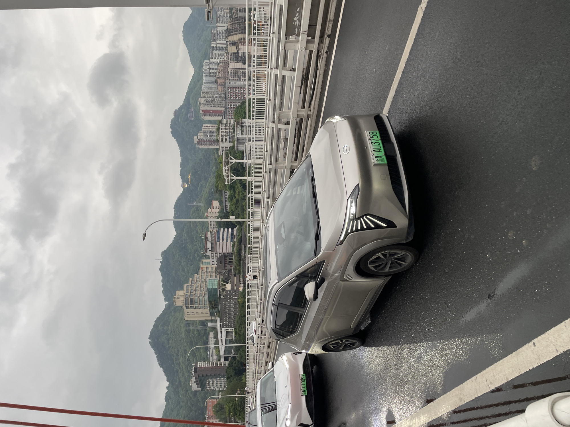 An overcast day in Nan'an District, China, features traffic on a bridge. In the foreground, a silver electric car with license plate "A AU3758" is driving in the right lane. To its left, a white electric car with license plate "A CT0097" is in the left lane. Both cars appear to be new models. The bridge has white railings, and a lamppost is visible in the background. Beyond the bridge, a densely populated city with apartment buildings and lush green hills under a cloudy sky is visible. The road surface is wet, indicating recent rain.