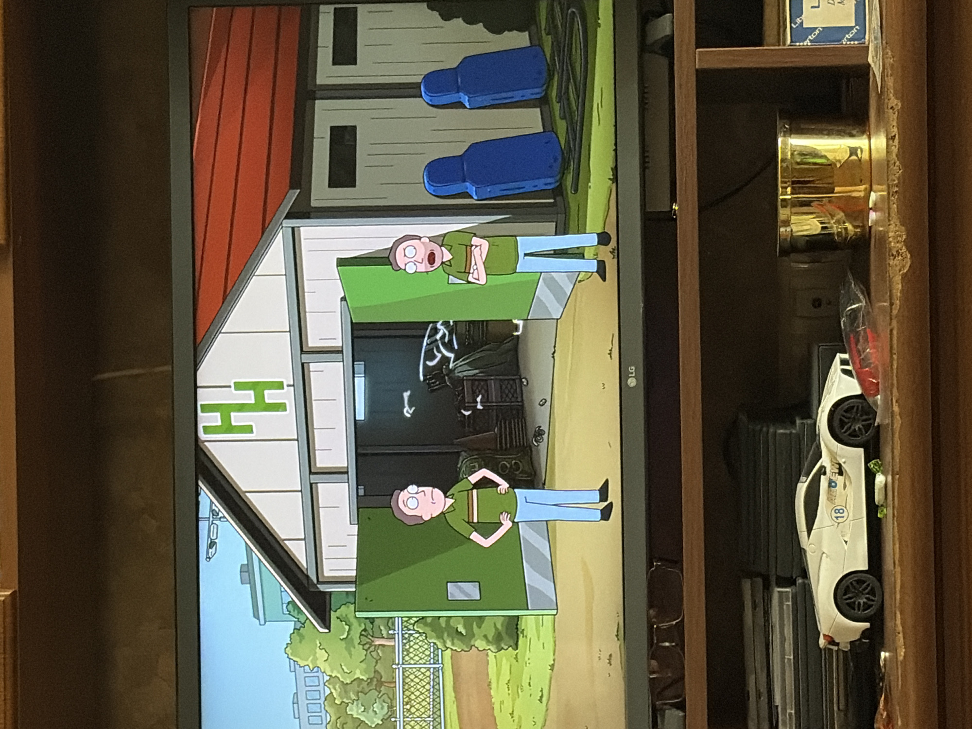 The image shows a television screen displaying an animated scene from the show "Rick and Morty." The scene depicts two identical male characters, Jerry Smith, standing in front of a garage with open green doors. The garage has a white facade with red siding on the roof and large green "HH" letters on the front. The character on the left has his hands on his hips, while the character on the right has his arms crossed. Inside the garage, there are several blue training dummies and various crates and equipment. The environment is grassy and appears to be outdoors, with a fence and trees visible in the background. Below the television, a shelf displays a white toy car and a gold container.