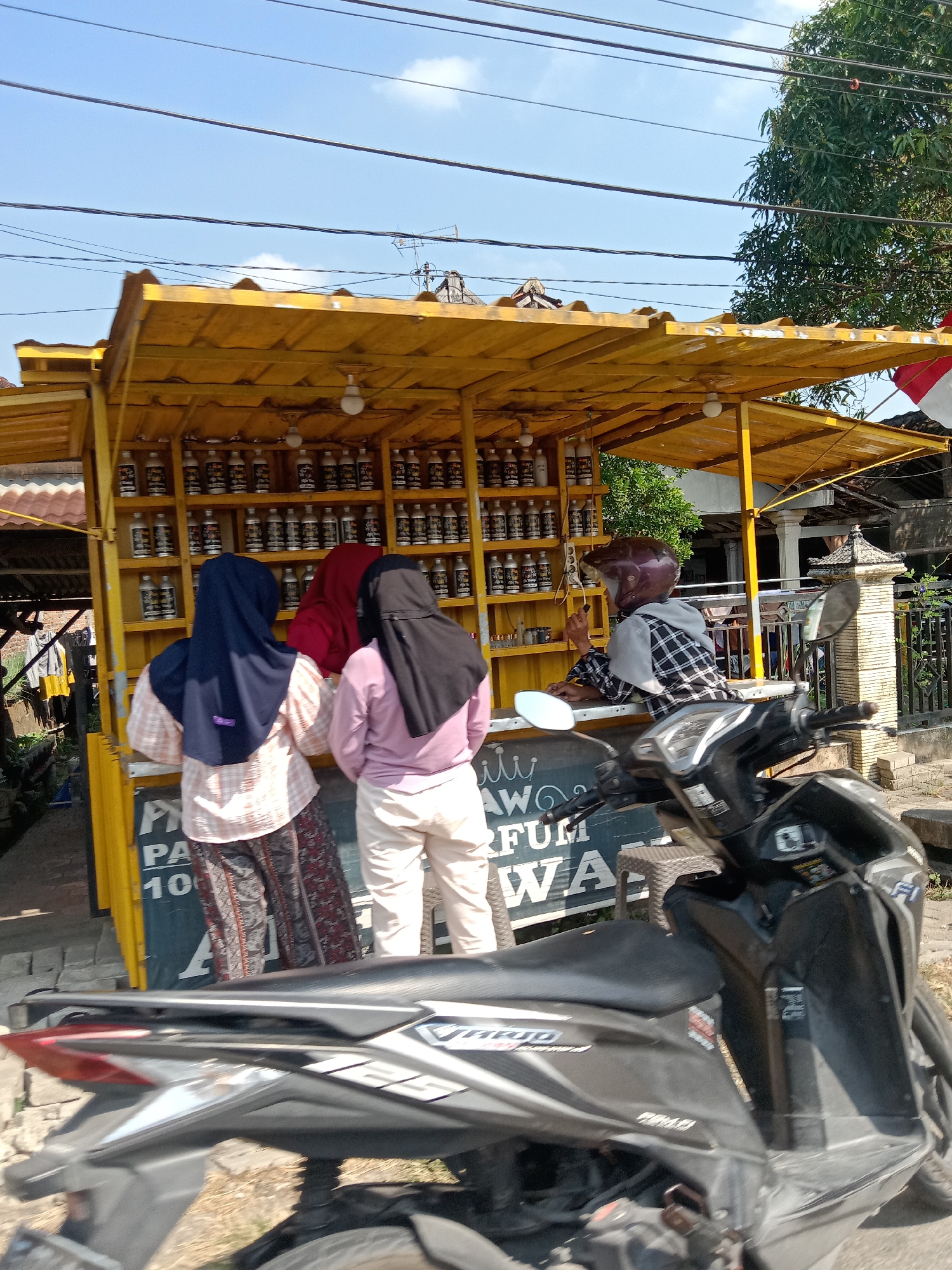 The image depicts an outdoor scene in Semawot, Indonesia, featuring a yellow open-air kiosk selling perfumes. Three individuals, two women and one person wearing a helmet, are present. The two women, dressed in hijabs and modest clothing, are standing in front of the kiosk, appearing to browse or purchase items. The person in the helmet, positioned behind the counter, seems to be interacting with them. The kiosk is stocked with numerous bottles, likely perfumes, arranged on shelves. A motorcycle is parked in the foreground, partially obscuring the kiosk. The background reveals a street scene with trees, buildings, and overhead wires, indicating a daytime setting. The overall atmosphere suggests a bustling local marketplace.