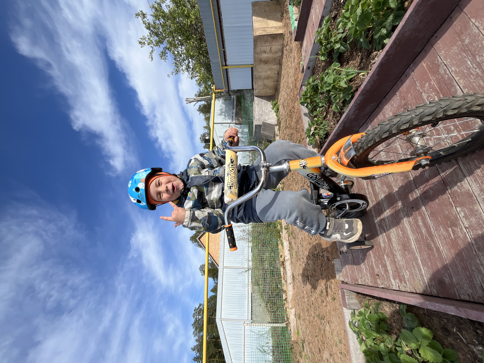 A young boy wearing a blue helmet and patterned jacket is riding a small, orange bicycle with training wheels on a wooden surface. He is looking at the camera with a playful expression and making a hand gesture. The background includes a blue sky with wispy clouds, trees, a greenhouse-like structure, and a raised garden bed with green plants. The location is Chaikovsky, Russia, based on the geotags and context clues.