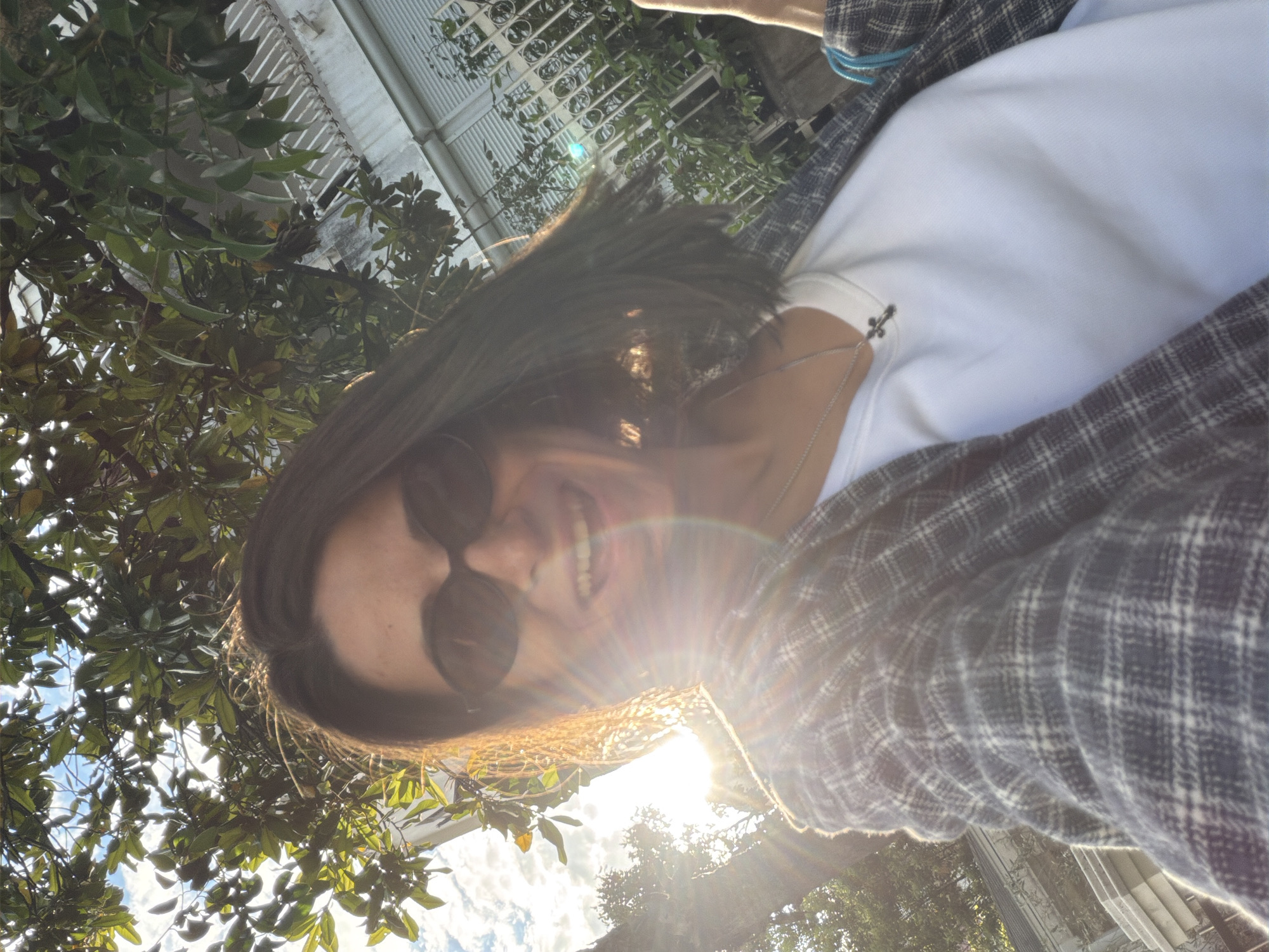 A person wearing sunglasses, a white shirt, and a plaid jacket is smiling in a photograph taken from a low angle, looking upwards. The sun is shining brightly, causing lens flare. Green leaves and branches are visible framing the subject, and a building with white railings is slightly visible in the background. The location is not identifiable as Budva, Montenegro, based on the visible details.