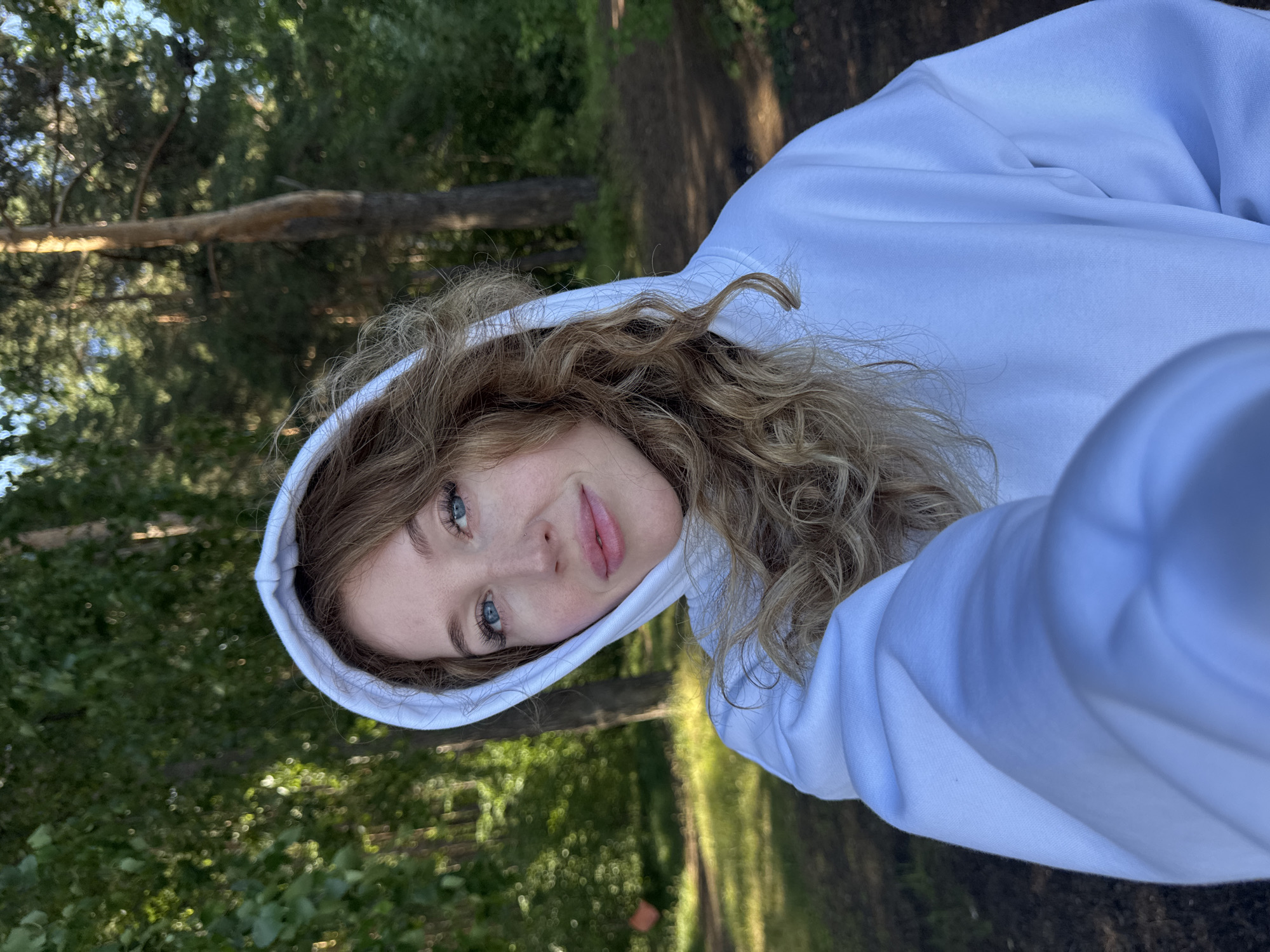 A young woman with blonde curly hair and blue eyes is taking a selfie in a forest. She is wearing a white hoodie and is looking at the camera with a slight smile. The background is a blurred forest scene with green trees and sunlight filtering through the leaves. The location is likely a park or natural area within the city of Chaikovsky, Russia, given the presence of pine trees and deciduous foliage common to the region. The lighting suggests it is daytime, possibly late afternoon due to the warm tones.