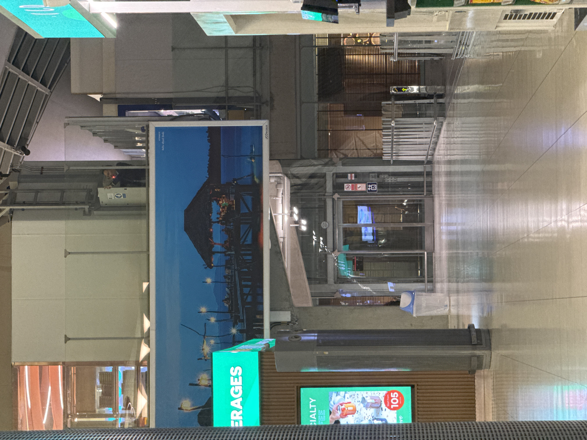 The image depicts an interior view of what appears to be a transportation hub or a commercial area in Nong Prue Subdistrict, Thailand. The scene is dominated by polished, reflective flooring that mirrors the overhead lighting and structural elements.
In the foreground, a tall, ribbed cylindrical object, possibly an air purifier or a decorative column, stands on the left. To the left of this column, a vertical digital display screen shows an advertisement for "CIALTY COFFEE," featuring images of cans and a price indicator of "105."
Further into the scene, a large horizontal banner displays a nighttime scene of a stilted structure over water with people seated, illuminated by lights, under a dark blue sky. The word "ERAGES" in bright green letters is visible on a screen to the left of this banner.
Behind the banner, glass doors lead into an interior space with closed wooden blinds. A person is visible through a glass partition on the upper level, standing near a control panel. Several metal barriers and railings are present in the midground. A blue and white trash receptacle is situated near the entrance.
On the right side of the image, another vertical digital display is partially visible, with a green illuminated section at the top. A white vertical structure with a grid pattern extends along the right edge of the frame.
The overall environment suggests a modern, well-lit public space, likely an airport, shopping mall, or entertainment complex. The lighting creates reflections that add depth to the image.