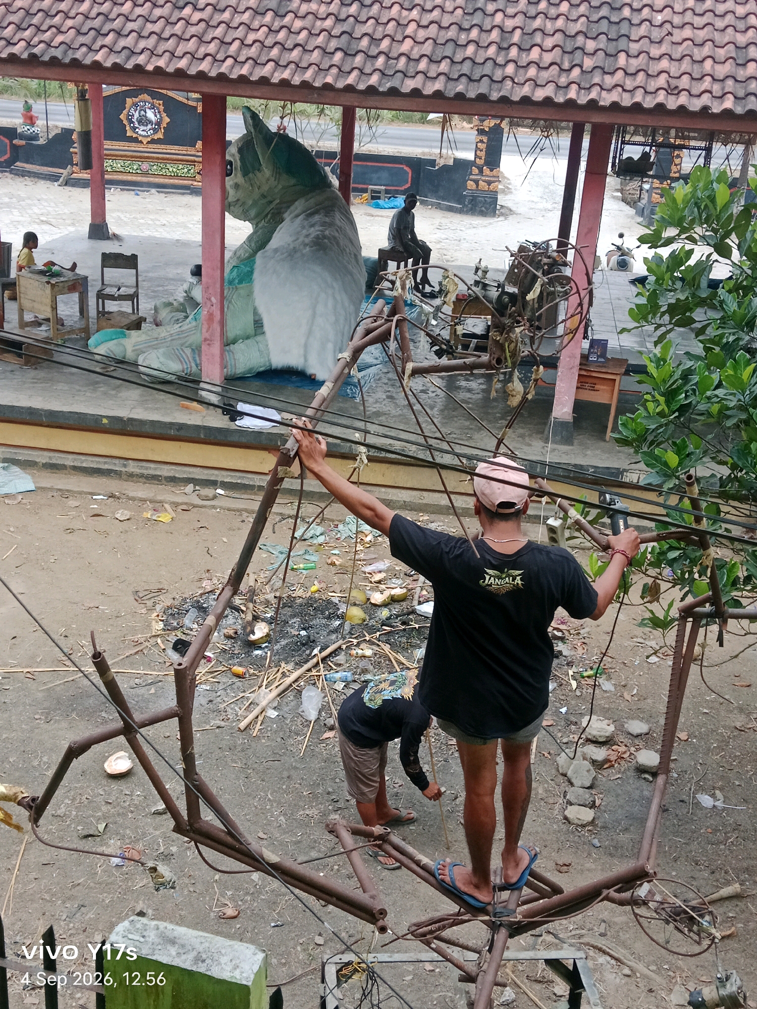 In Rama Dewa, Indonesia, a person stands precariously on a metal structure, holding onto electrical wires. The person is wearing a black t-shirt with the word "JANGALA" on the back and a pink cap. Another person, wearing a long-sleeved black shirt and tan shorts, is visible lower down on the structure, holding a stick. The ground below is littered with debris, including what appears to be charred material, coconuts, and plastic bottles. In the background, a large, inflatable cat sculpture is visible under a covered structure, with a child sitting at a small table nearby. The scene suggests a candid moment captured amidst what appears to be a maintenance or construction activity near a local gathering space.