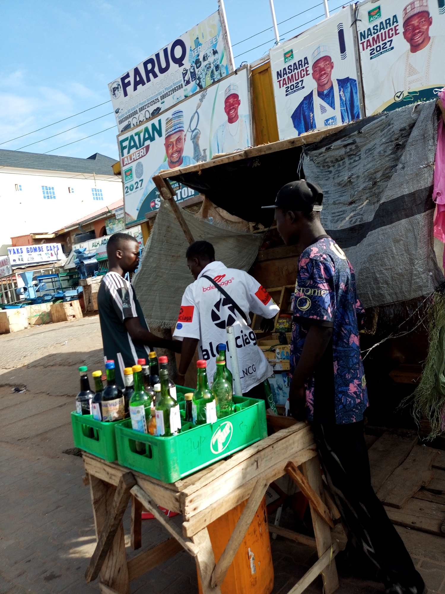 A man in a black cap and a patterned shirt stands with his back to the camera, looking at another man in a green and black striped shirt who is looking at a display of bottles on a wooden table. The man in the striped shirt is wearing a black cap. In the background, there are several large campaign posters with images of men in traditional attire. One poster reads "FARUQ COMMUNICATION". Another poster has the name "NASARA TAMUCE 2027" and "ADC" logo. The scene appears to be a street market in Arawa, Nigeria, with the posters suggesting an upcoming election or political campaign. The bottles are displayed in green plastic crates on a rustic wooden table. The ground is paved. The sky is blue with some clouds.