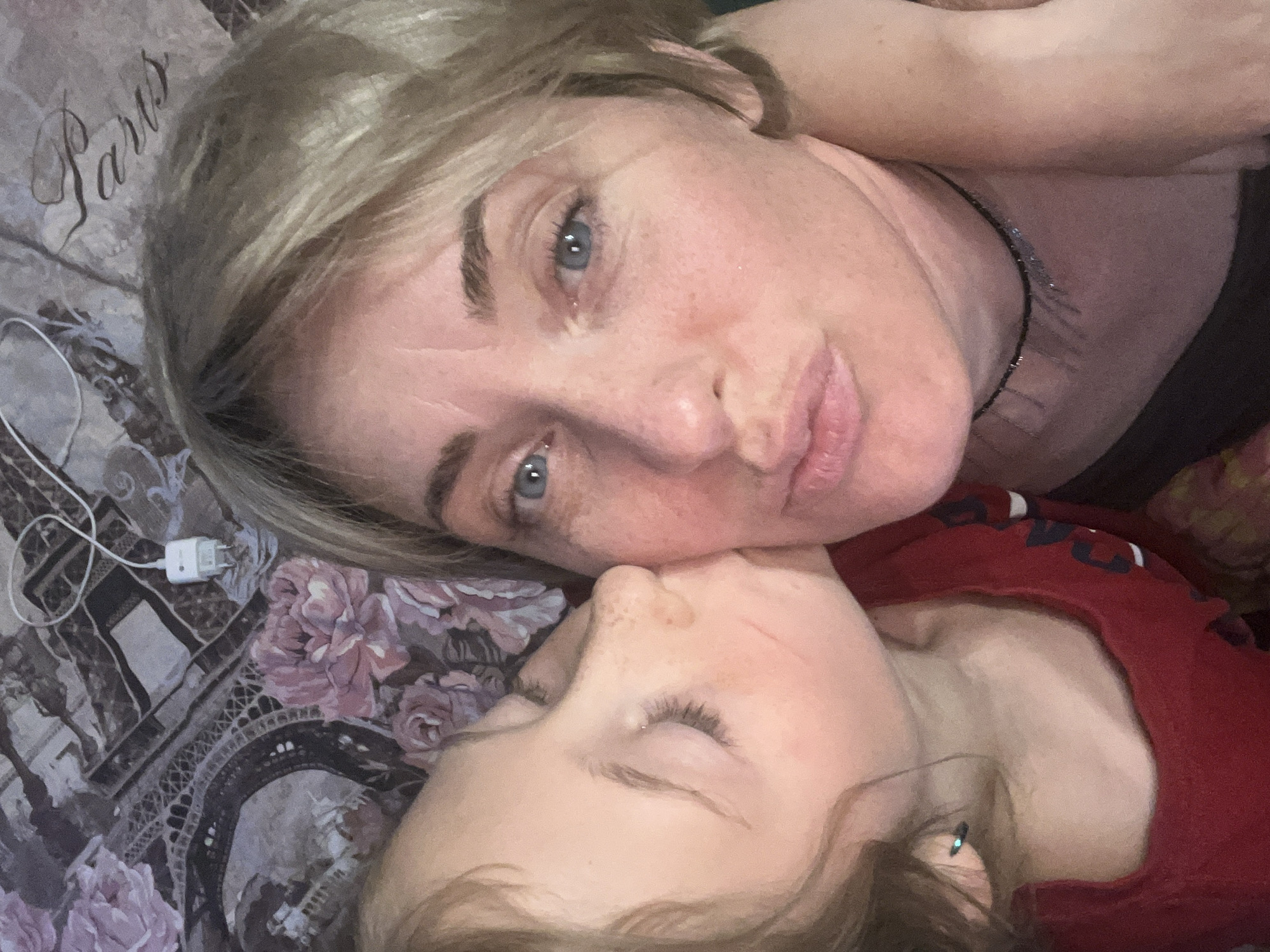 A close-up, overhead shot shows a woman with blonde hair and blue eyes being kissed on the cheek by a child. The woman has a necklace on and is wearing a dark top. The child is wearing a red shirt. They are lying on a bed with a patterned comforter depicting Paris landmarks and flowers. A white phone charger is visible on the comforter. The image is tightly cropped, with the faces of both individuals prominently featured. The setting appears to be indoors, likely a bedroom. There is no information to suggest the image is located in Saratov, Russia.