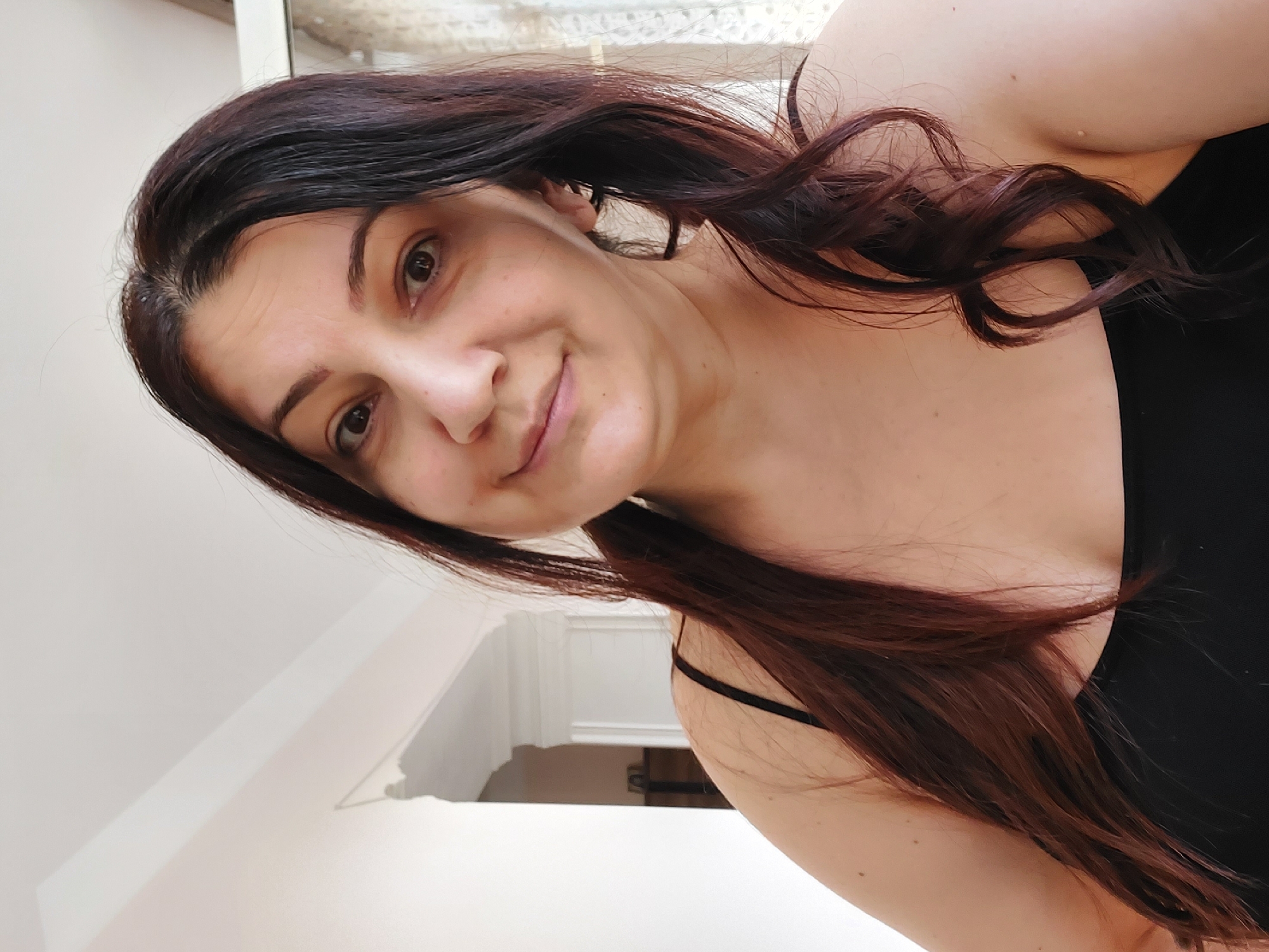 A medium shot selfie of a woman with long, dark brown hair styled in two low pigtails. She is wearing a black tank top. The woman has brown eyes, arched eyebrows, and a slight smile. She is positioned in front of a white wall with decorative molding. The background suggests an indoor setting, possibly a home, with hints of a window with sheer curtains on the right side and a doorway or archway on the left. The lighting appears natural, coming from the front and possibly from the right. The image is taken from a slightly upward angle. There is no visible indication of the city of Yerevan, Armenia within the image itself.