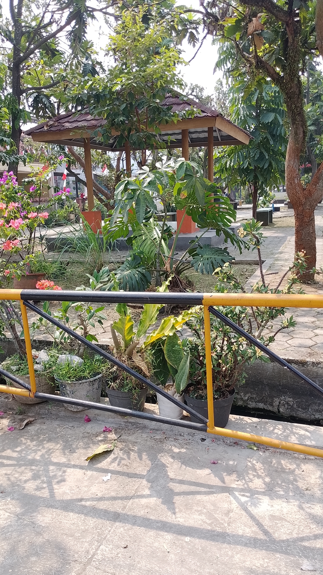 The image captures an outdoor scene in Bojongsoang, Indonesia, featuring a gazebo in the background surrounded by lush greenery and potted plants. A yellow and black railing runs across the foreground, obscuring a canal or drainage ditch. The ground is paved with concrete, and shadows from the surrounding trees are cast upon it. The overall atmosphere suggests a park or a public garden space. No people are visible in the image.
