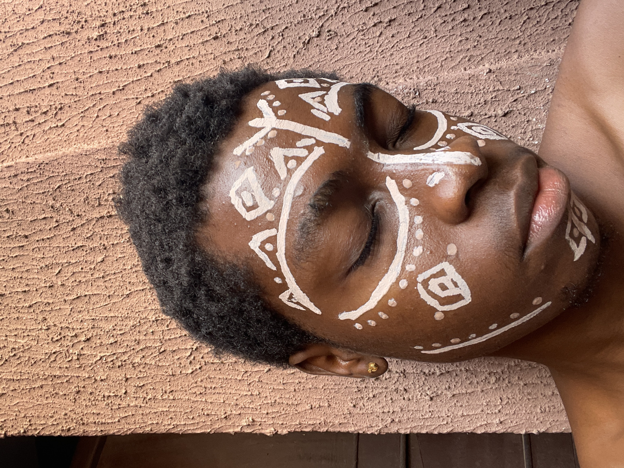 A close-up, vertically oriented image depicts a person with dark skin and afro-textured hair, lying down. White tribal-like markings are painted on the person's face. The person's eyes are closed, and their face is in profile against a textured, light-brown surface. A small gold earring is visible in their ear. Part of the person's shoulder and neck are visible in the lower right. The location, Nkpor, Nigeria, cannot be verified from visual details in the image.