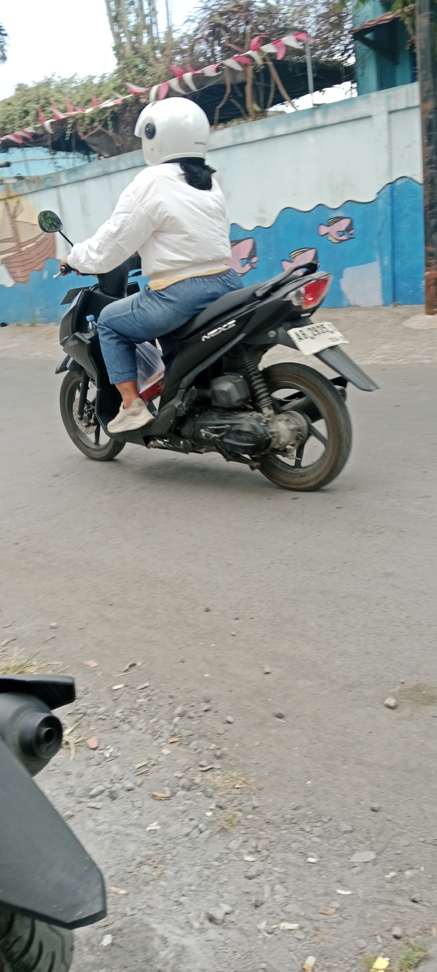 A person wearing a white helmet and a white long-sleeved shirt is riding a black scooter on a paved road. The person is also wearing light blue jeans and light-colored sneakers. The scooter has a license plate that reads "AB 2333 DJ". In the background, there is a wall with a mural of a ship on the left and fish on the right. The wall is blue and white. There are trees and foliage visible above the wall. The road surface appears to be asphalt with some gravel and debris. The image was taken in Purwomartani, Indonesia.