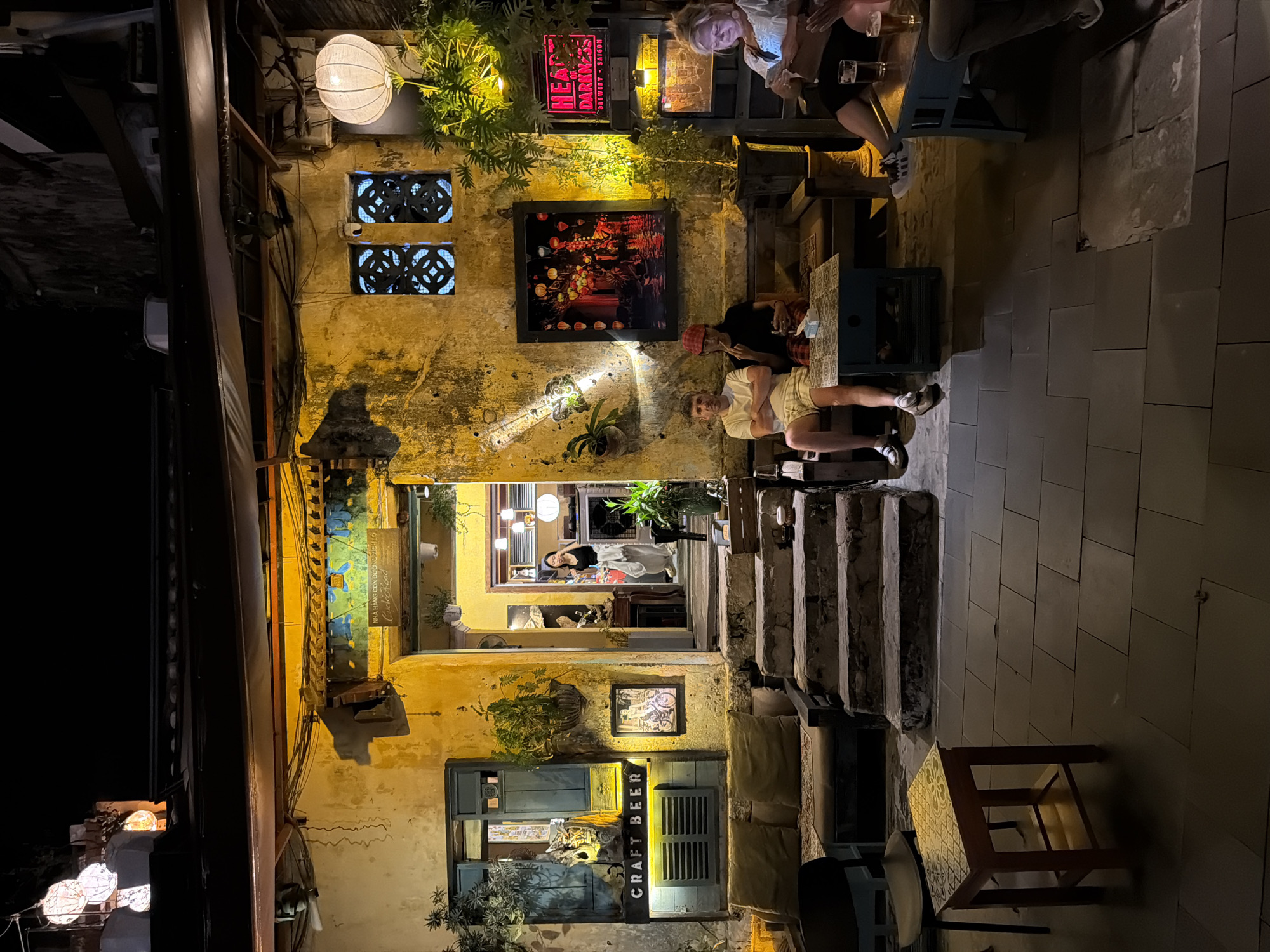 The image shows the exterior of a bar or restaurant in Hội An Ward, Vietnam, at night. The building has a weathered, yellow facade with exposed brick and peeling plaster. A sign above a doorway reads "CRAFT BEER" in illuminated lettering. To the right of the entrance, a woman stands in the doorway. In front of the building, several people are seated at tables and on a bench. A man in a white t-shirt and shorts sits on a bench, and next to him, another person wearing a red hat is smoking. Further to the right, a woman is looking at her phone while seated at a table with drinks. The area is paved with stone tiles, and plants are visible in pots around the establishment. Lanterns hang from the building's overhang, and a round, white lantern is illuminated on the right side of the facade. A framed artwork depicting lanterns is hung on the wall.
