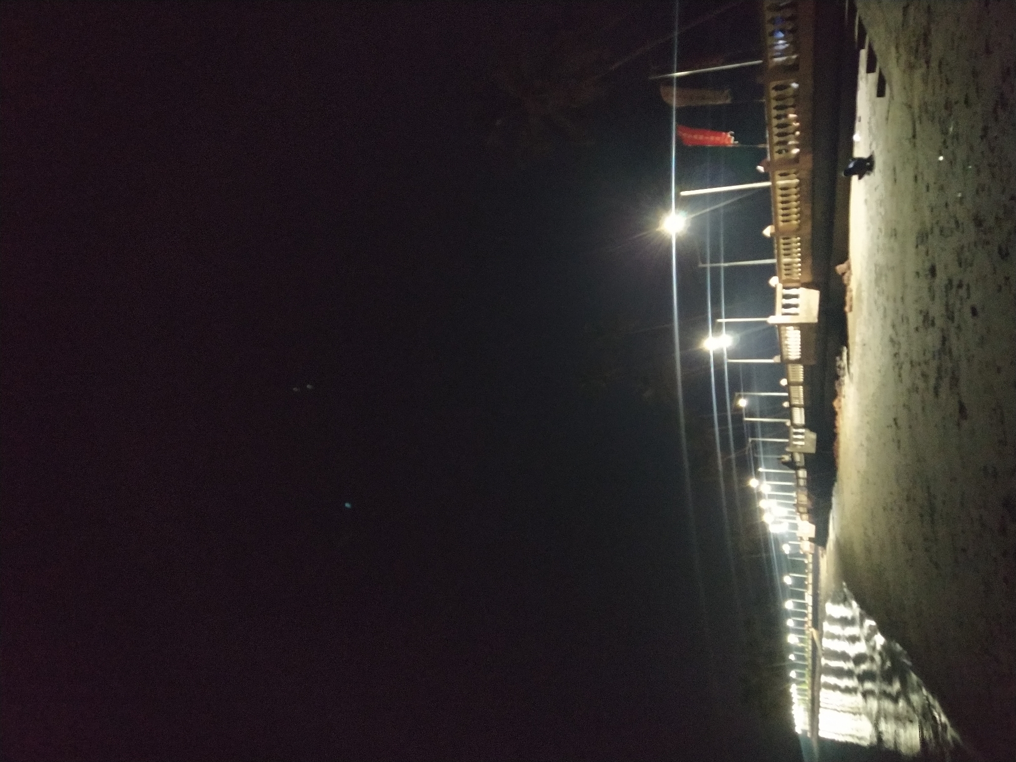 The image depicts a nighttime scene along what appears to be a waterfront promenade or walkway in Badem, India. A row of evenly spaced streetlights illuminates the walkway, casting reflections on the wet surface below, possibly water or wet sand. The lights reveal a retaining wall or edge of the walkway to the right. The left portion of the image is dominated by darkness, suggesting a view toward the water or an unlit area. A red cloth is hanging from above on the right side.