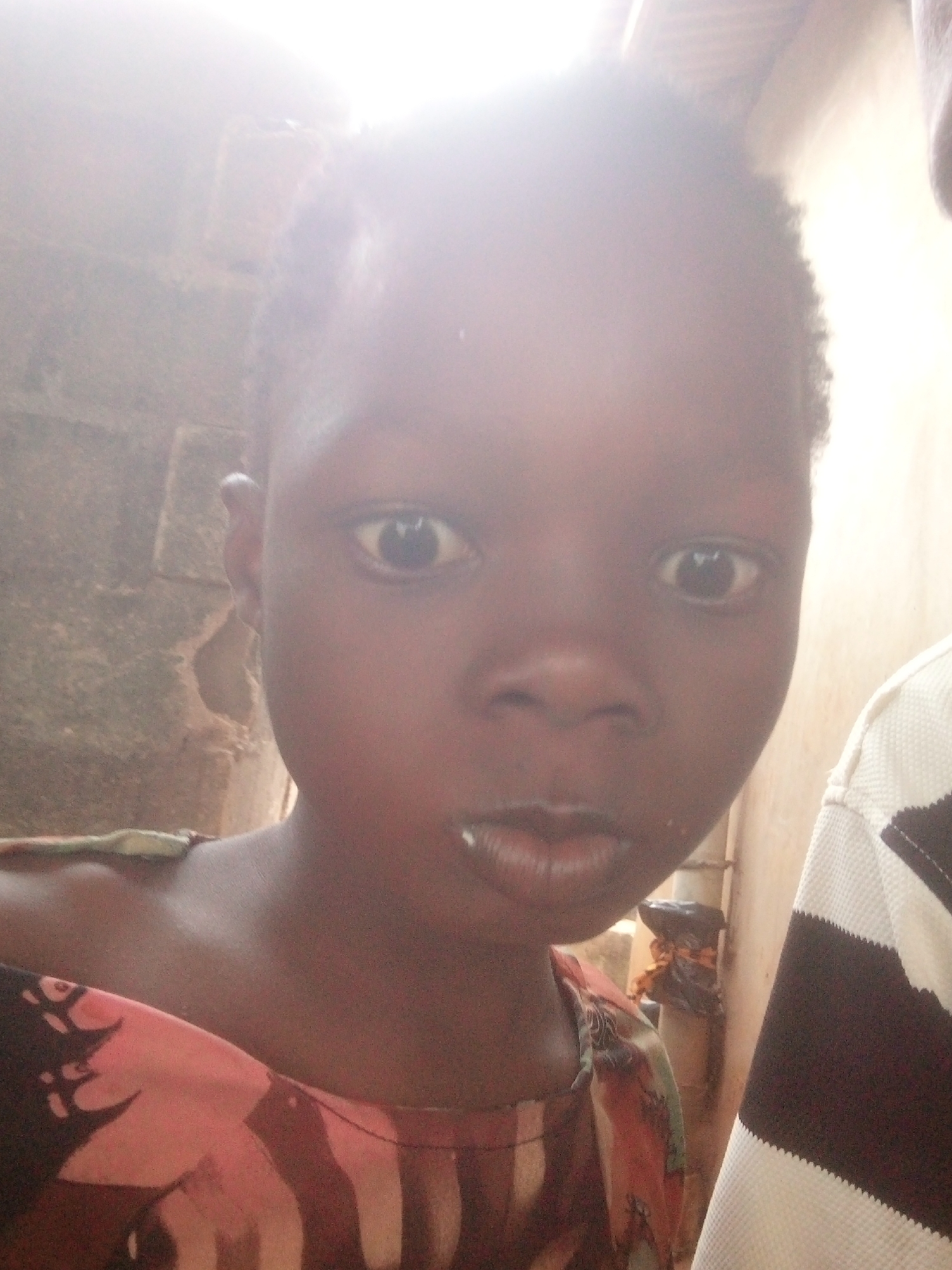 A young, dark-skinned child is the primary subject of this close-up image. The child has short, dark hair and is looking directly at the camera with a slightly pursed mouth. Traces of something white are visible near the mouth. The child is wearing a garment with a print in reddish and darker tones.
To the left of the child, a rough, textured wall is partially visible. To the right, a person wearing a striped black and white shirt is partially in the frame. Behind the child, in the background, there's a pipe or pole with what appears to be a black bag or object attached. The lighting is uneven, with bright areas obscuring details, particularly near the top of the image. The location is Unknown, Unknown.