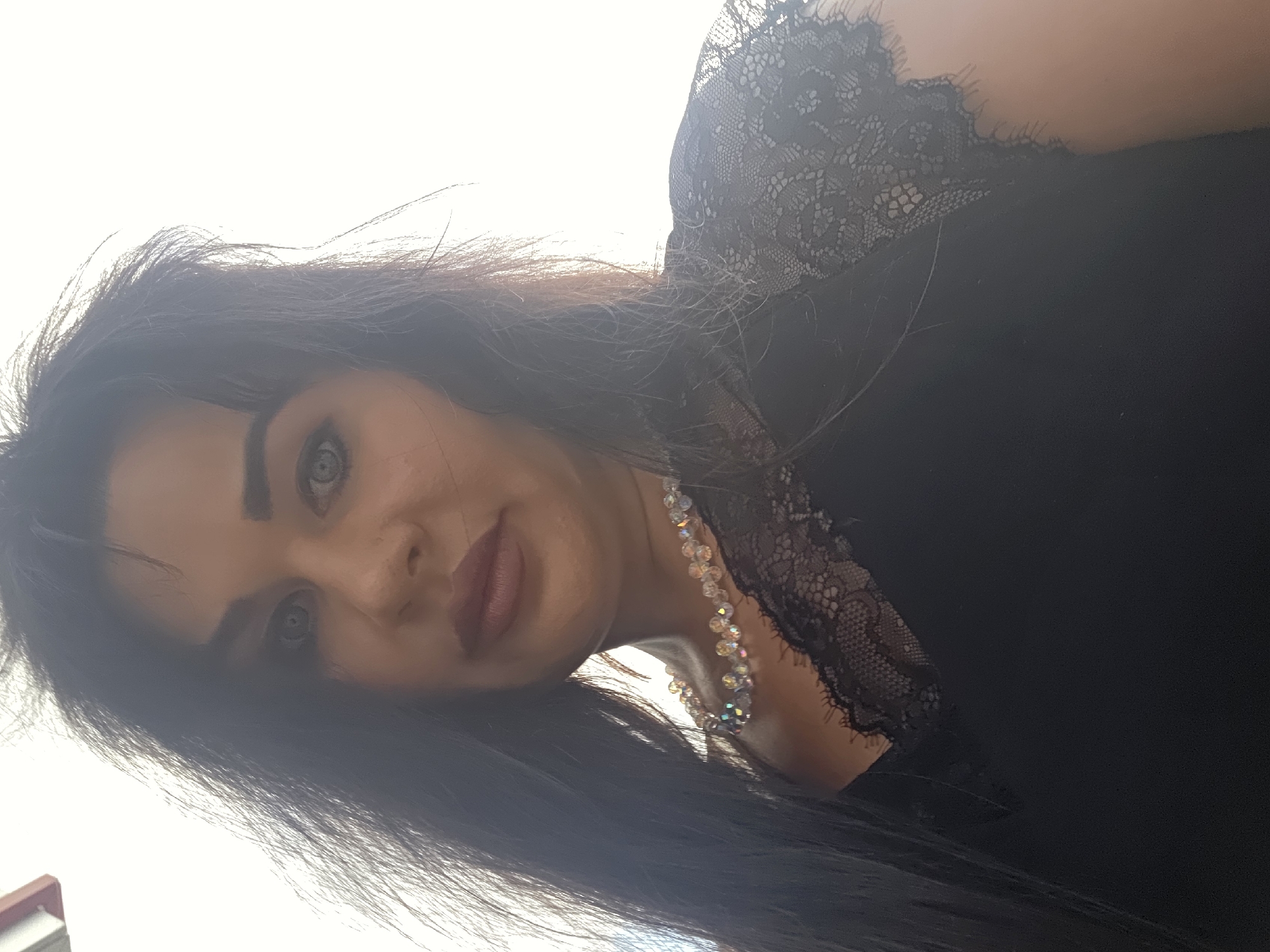 A woman with long, dark, wavy hair is looking at the camera. She has fair skin, light-colored eyes, and dark eyebrows. She is wearing a black top with lace trim on the sleeves and neckline. She is also wearing a necklace made of clear, iridescent beads. The background is a bright, blown-out sky, suggesting an outdoor setting. The image was taken in Sochi, Russia.