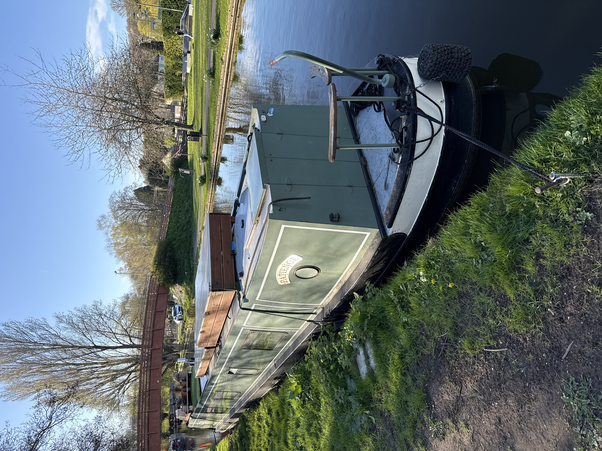 A docked narrowboat, named "Patience", is moored along a canal in Dacorum, United Kingdom. The vessel is green with white trim and a black fender. A rope connects the boat to the grassy bank. The canal itself appears dark and still. Beyond the canal bank, there is a grassy area, trees, and a footbridge. The sky is blue with scattered clouds.