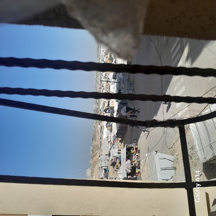 An outdoor scene viewed through a barred window captures a dusty, sunlit cityscape. Makeshift shelters and small buildings cluster along a road with pedestrian crossings. People are visible in the scene. The sky is clear and blue. A telephone pole is also visible. The bars are dark and somewhat rusty.