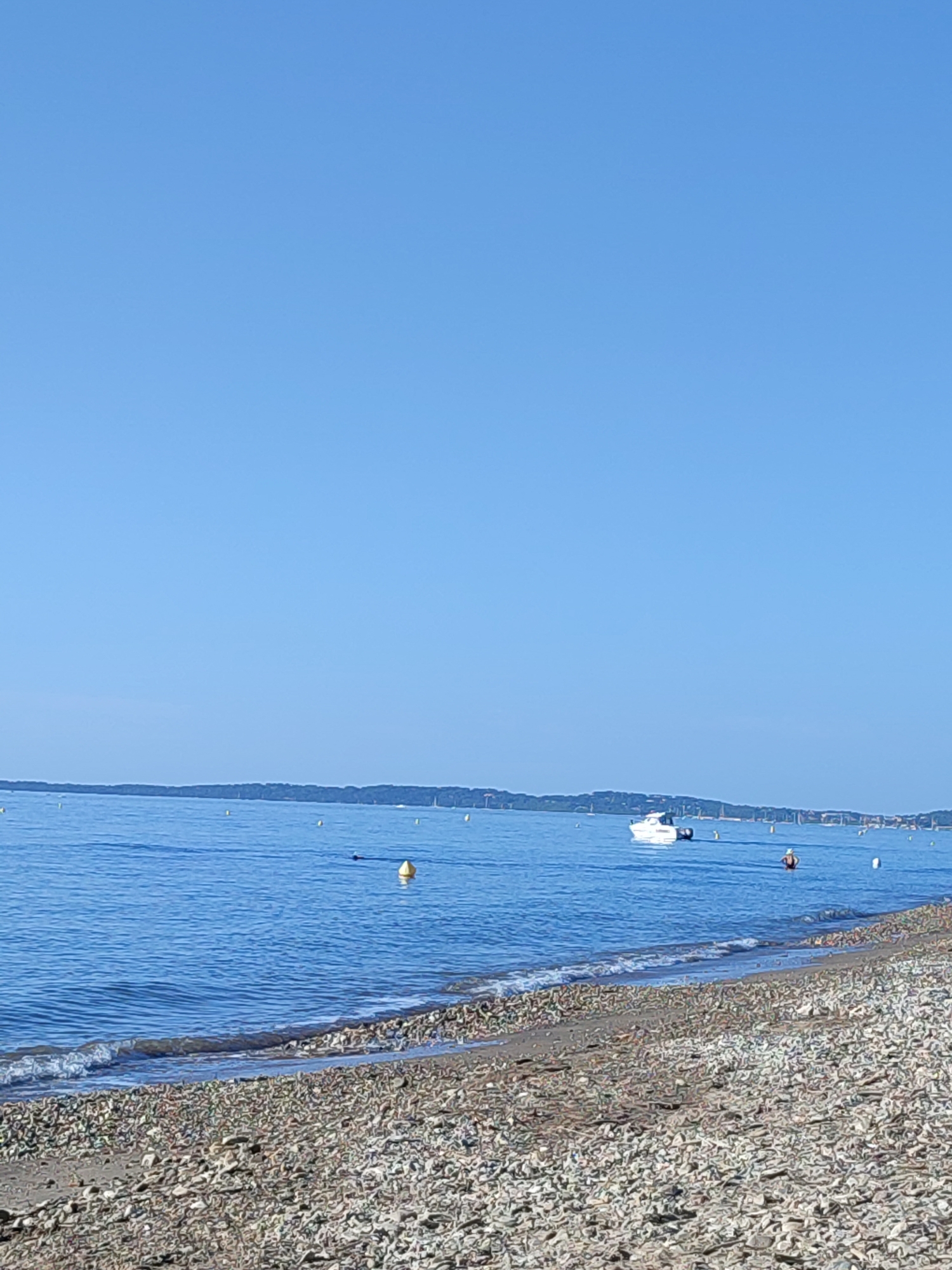 A wide shot captures a tranquil beach scene in Hyères, France, under a clear blue sky. The foreground is a pebble beach, with the stones varying in size and color, giving it a textured appearance. Gentle waves lap at the shore, creating a thin line of white foam. The sea stretches out to the horizon, a calm expanse of blue water. In the middle distance, a white motorboat with a dark dinghy tied to its stern is anchored. Several yellow buoys are visible scattered across the water, marking an area. Further in the distance, a long, tree-lined landmass forms the horizon, suggesting a coastline or island. A person can be seen in the water near the boat, appearing to be swimming or wading. The overall impression is one of peace and natural beauty on a sunny day.
