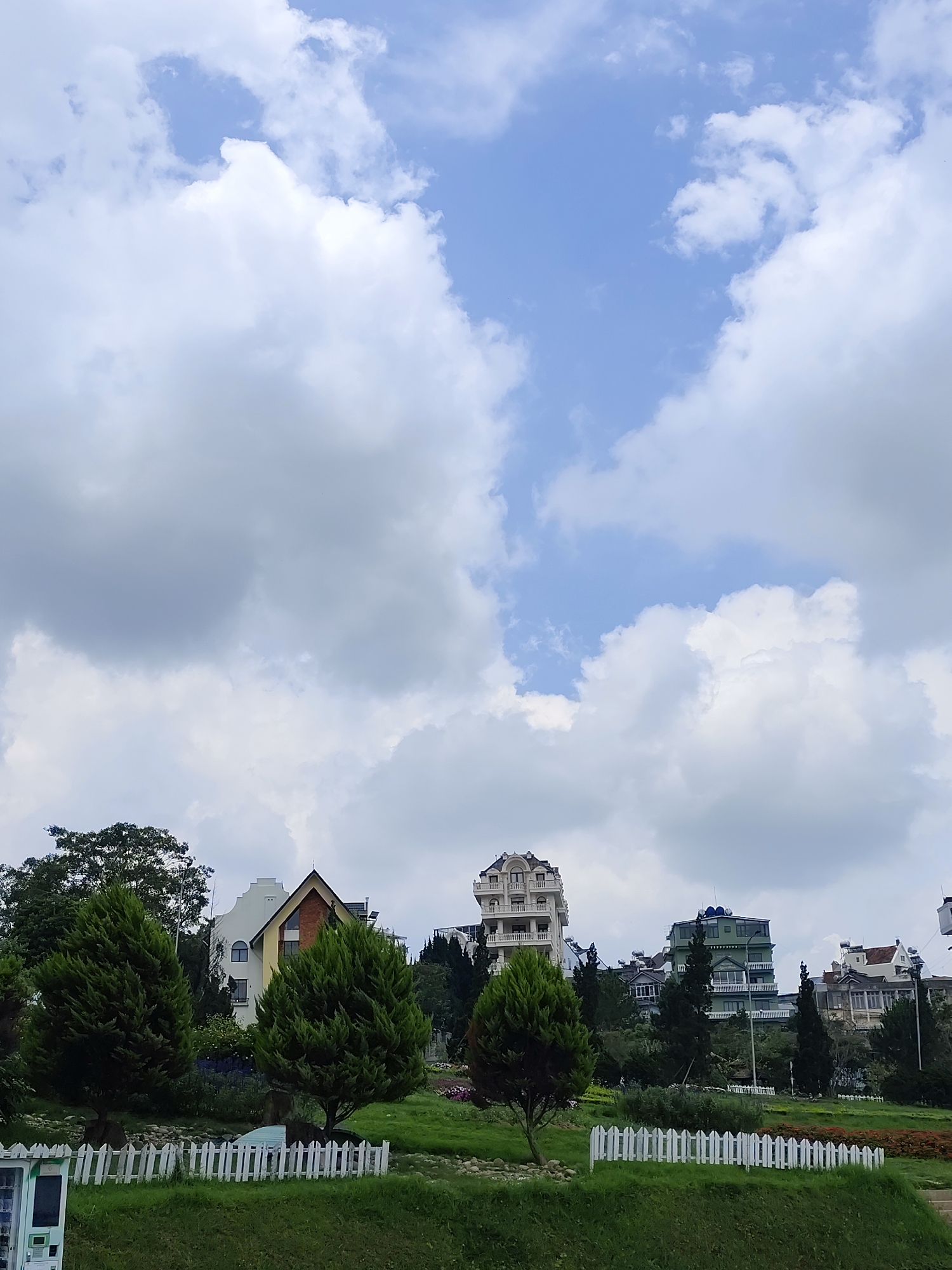 The scene shows a picturesque landscape in Da Lat, Vietnam, dominated by a sky filled with cumulus clouds. Below, a green lawn extends, partially bordered by a white picket fence. Scattered across the landscape are evergreen trees, intermixed with various buildings. Among the buildings are structures with distinctive architectural styles, including one with a pointed roof and another resembling a multi-story apartment complex. The composition includes details like a vending machine near the fence and glimpses of flowers, adding to the overall charm of the scene.
