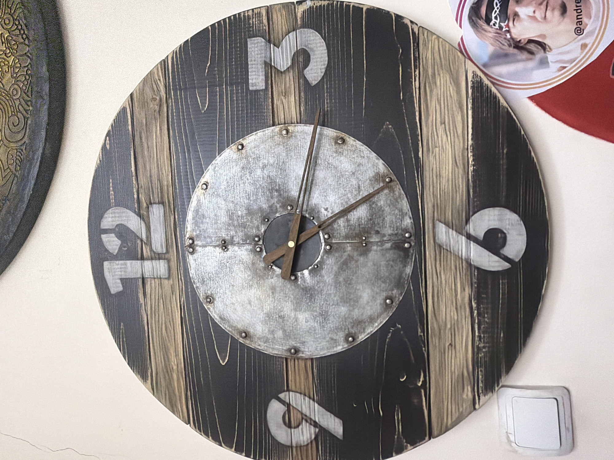 A wooden clock with a metal face and Roman numerals is mounted on a textured wall. The clock has dark wood planks with lighter wood accents. The numbers 12, 9, 3, and 6 are visible. The clock hands are made of dark wood. A white light switch is visible on the wall to the left of the clock. A circular item with a picture of a man is in the lower right corner, partially overlapping the clock. The man in the photo has a bandana on his forehead with a star design and is wearing a white t-shirt. The text "@andre" is visible below the man's image. The scene appears to be indoors. The location is identified as Maloyaroslavets, Russia.