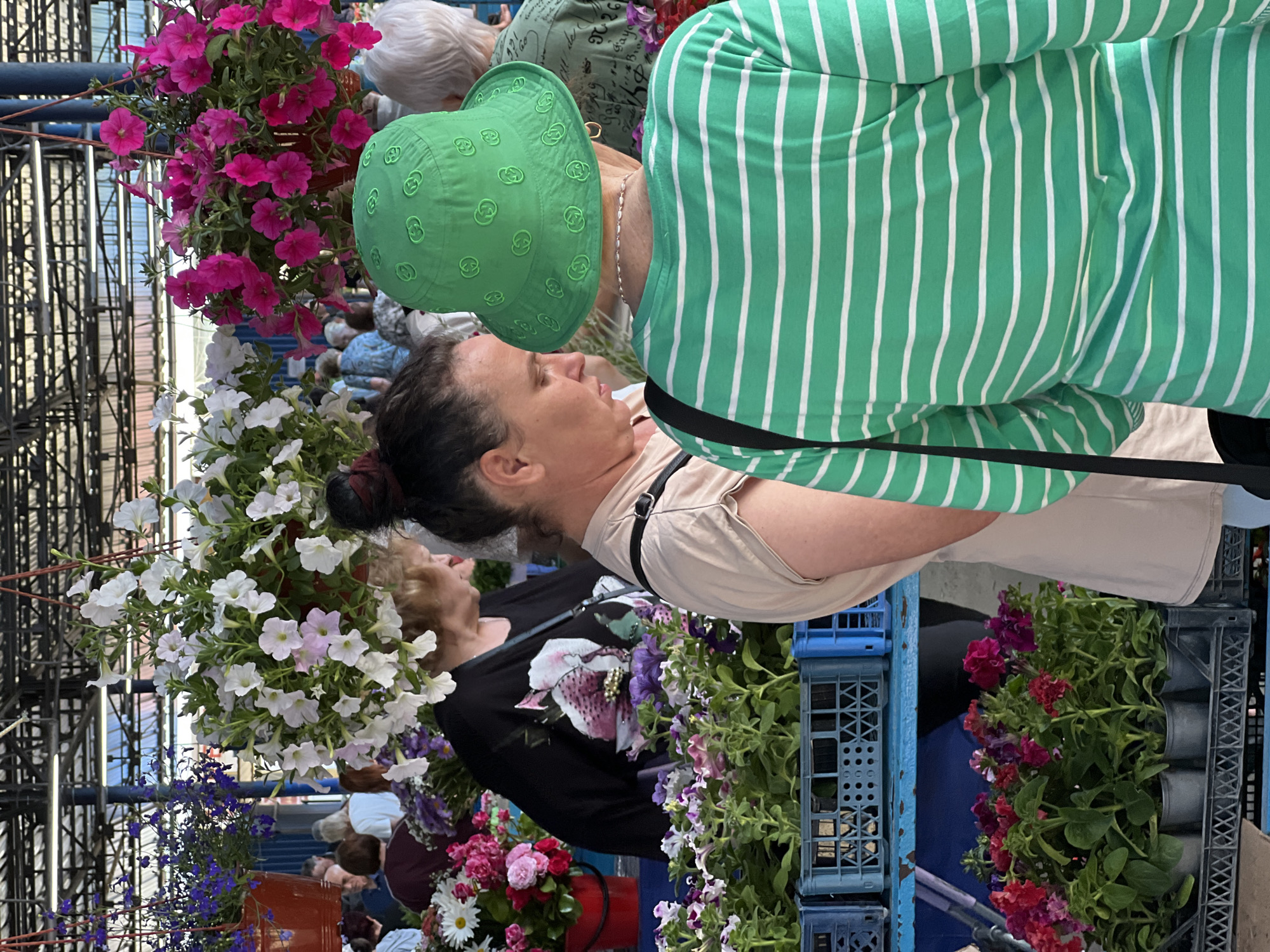 The image captures a vibrant outdoor flower market in the city of Krasnodar, Russia, under a large, light-colored corrugated roof supported by a metal framework. The scene is populated by several individuals, predominantly women, engaged in browsing or selling floral products.
In the immediate foreground, a person is seen from behind, wearing a bright green bucket hat embossed with repeating "G" logos and a green and white horizontally striped long-sleeved shirt. A silver chain necklace is visible around their neck. To their left, a woman with dark hair tied back in a bun is partially visible in profile, wearing a plain beige short-sleeved top, looking downwards.
Further to the left, an older woman with blonde hair, wearing a black short-sleeved shirt adorned with a prominent pink and white floral design, is observed looking towards the right, seemingly interacting with market goods.
The market stalls display an abundance of flowers. At the lower left, blue plastic crates hold numerous small potted flowering plants, predominantly petunias in shades of purple, pink, and white. A red bucket contains a bouquet of cut flowers, including white daisies and pink roses. Overhead, three large hanging baskets are prominently featured: one on the left with purple lobelia in an orange pot, a central basket overflowing with white petunias, and another on the right featuring bright fuchsia petunias.
In the background, numerous additional market-goers, mostly adults, are visible, moving and observing the various stalls, though their features are indistinct due to distance and blur. The environment suggests a lively, organized outdoor marketplace dedicated to horticulture.