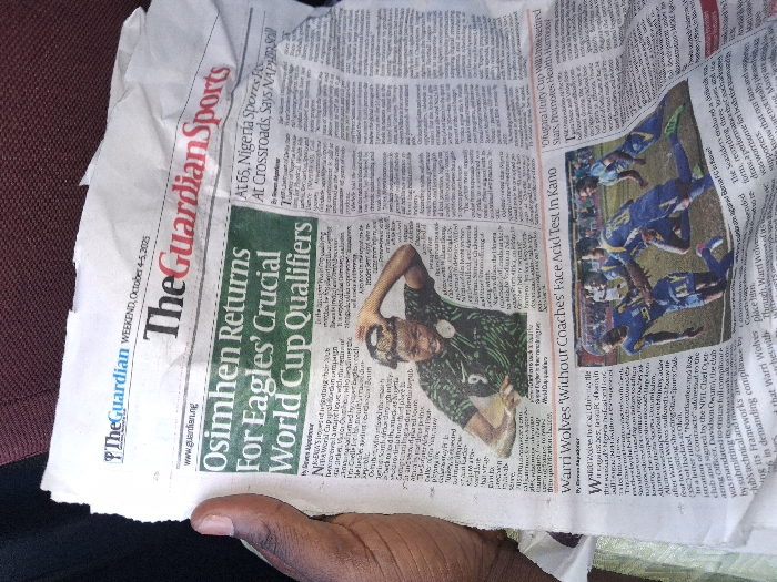 A crumpled, off-white copy of "The Guardian Sports" newspaper, dated "WEEKEND, October 4-5, 2025", is held by a dark-skinned hand on the left side of the frame. The newspaper's primary headline, highlighted in green, reads "Osimhen Returns For Eagles' Crucial World Cup Qualifiers". Below this, a photograph depicts a male soccer player, likely Victor Osimhen, wearing a dark green Nigeria national team jersey with the number "9" visible, his hands near his head. To the bottom-center, an article titled "Warri Wolves 'Without Coaches' Face Acid Test In Kano" accompanies a photograph of multiple male soccer players in blue and yellow uniforms actively engaged in a match on a green field. The background consists of a blurred, reddish-brown surface, possibly upholstery. This scene is observed within the city of Kungana, Nigeria.