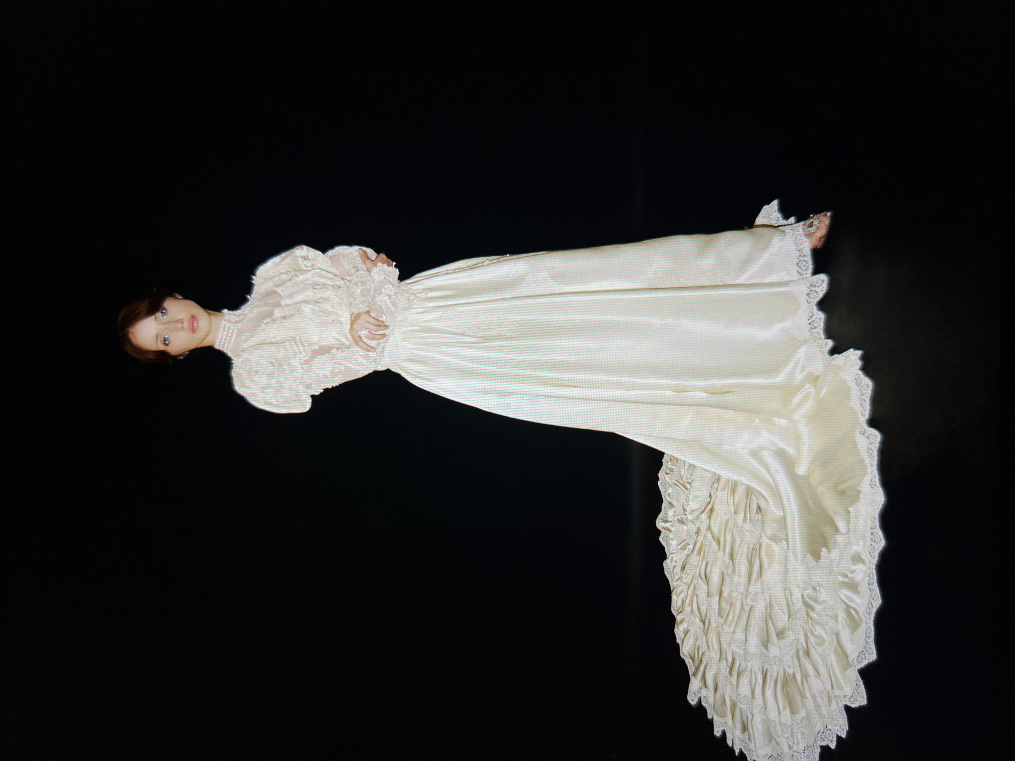 A young woman stands in a white, vintage-style wedding dress with a long train. The dress features puff sleeves, lace detailing on the bodice and cuffs, and a high neckline. Her arms are crossed, and she is looking directly at the camera with a neutral expression. The background is solid black. The train of the dress is spread out on the floor, decorated with ruffles and lace trim. She is wearing jeweled heels. The lighting is a studio-style light, casting a subtle shadow behind her. The image is a full-body shot, with the woman positioned slightly off-center to the right. The photograph is taken from a slightly elevated angle.