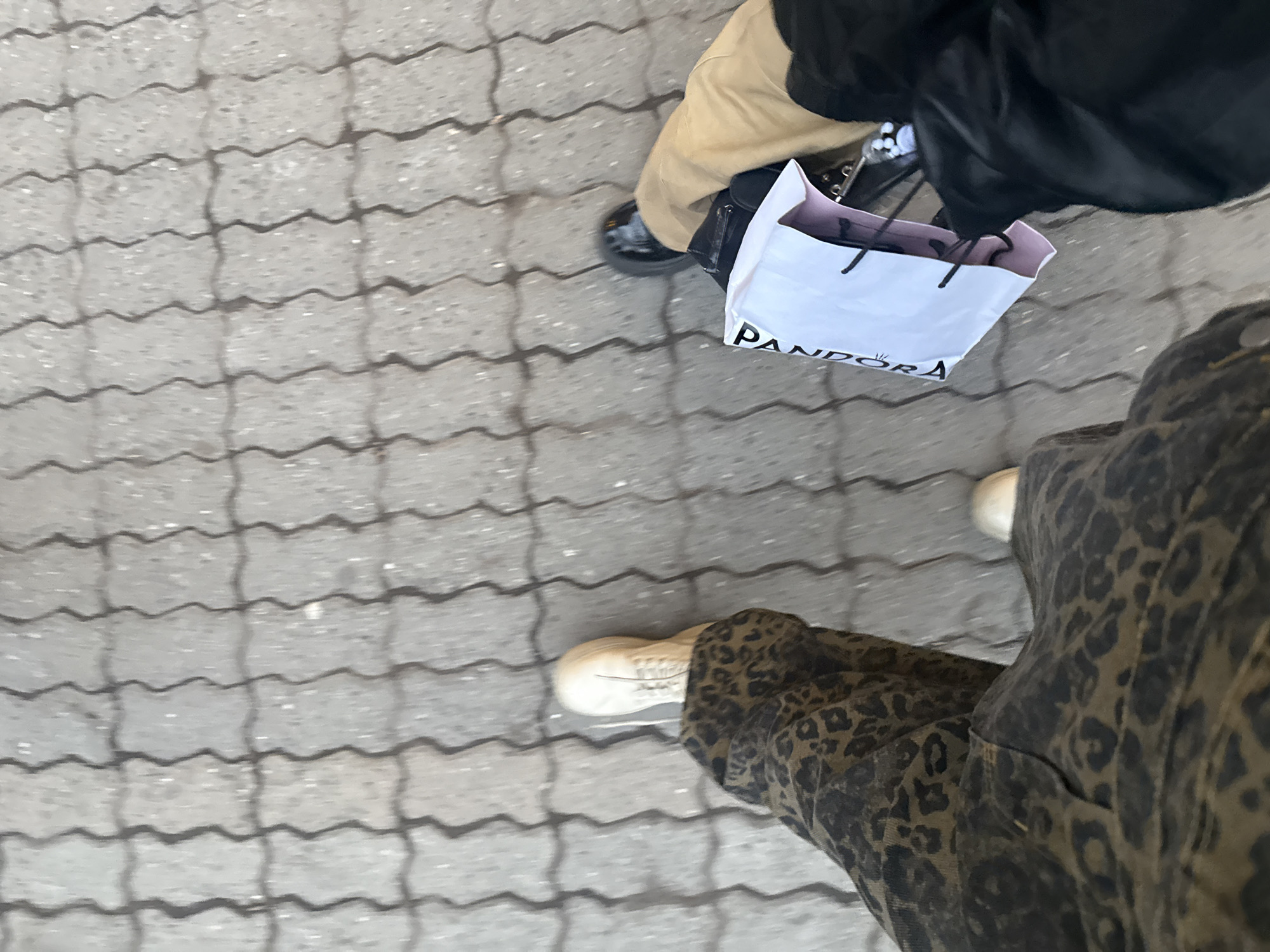 The scene captures a low-angle view of two individuals walking on an interlocking paver sidewalk in Yerevan, Armenia. The light gray pavement exhibits a wave-like pattern delineated by dark gray grout. The individual on the left wears brown and black leopard print trousers and light-colored athletic shoes, with the right foot extended forward. The individual on the right wears black trousers, a tan outer garment, and black flat shoes. This individual carries a white shopping bag, partially displaying "PANDORA" in black lettering, and a black handbag. Both individuals are in motion, walking forward.