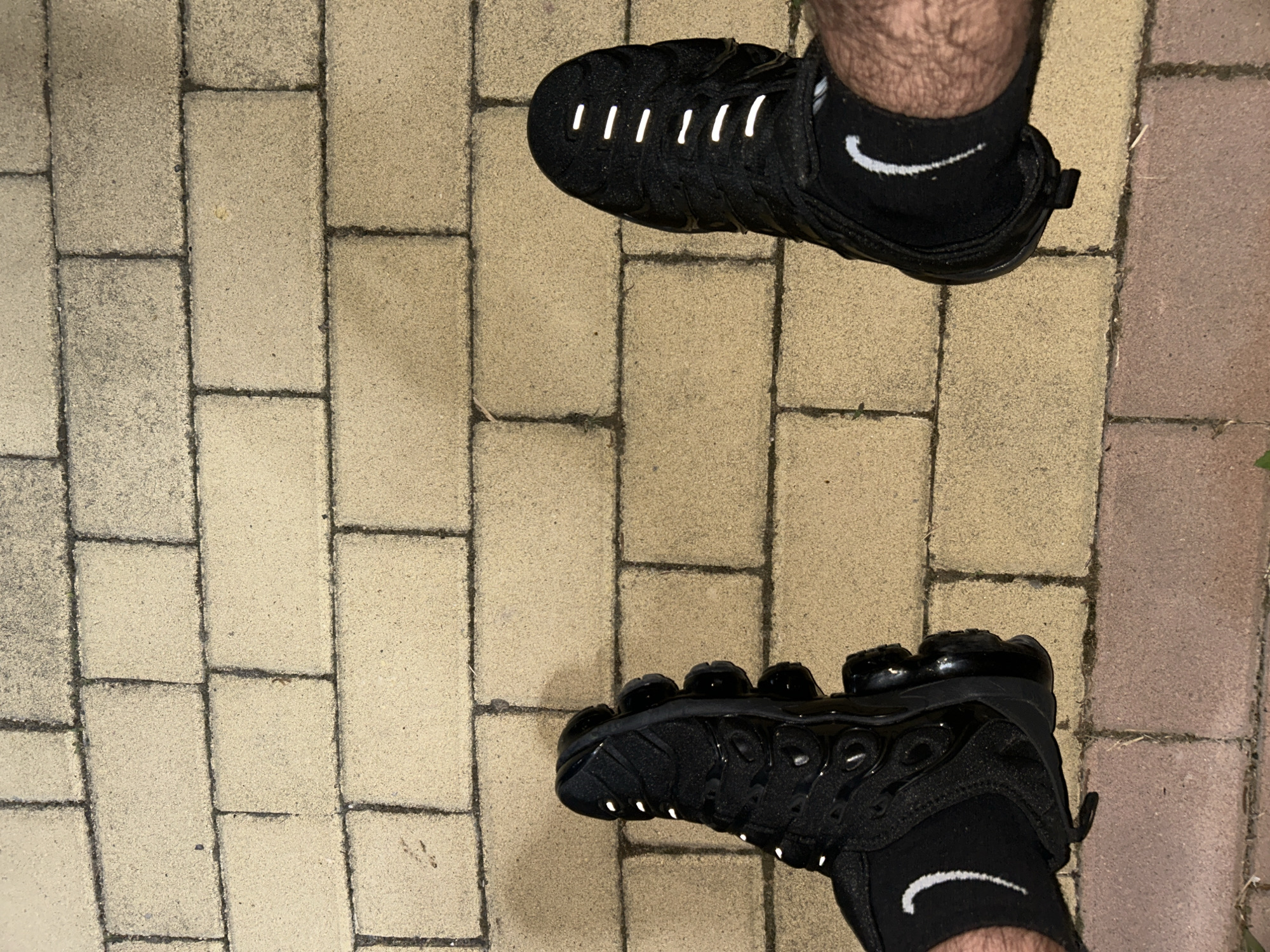 A top-down photographic view depicts two human feet, clad in black socks and black Nike Vapormax Plus sneakers, resting on a light-colored, rectangular brick paved surface in the city of Dzhebel, Bulgaria. The paving comprises individual bricks arranged in a consistent pattern with dark grey grout lines. Visible portions of the lower legs exhibit hair. Both feet are adorned with black ankle socks featuring a white Nike swoosh logo positioned on the outer side of each ankle. The left sneaker is oriented to reveal its distinctive side profile, including the segmented clear sole and a textured upper. The right sneaker is viewed from directly above, showcasing several parallel, reflective white strips across the instep of the shoe. The scene is illuminated by an ambient light source, accentuating the reflective elements on the footwear.