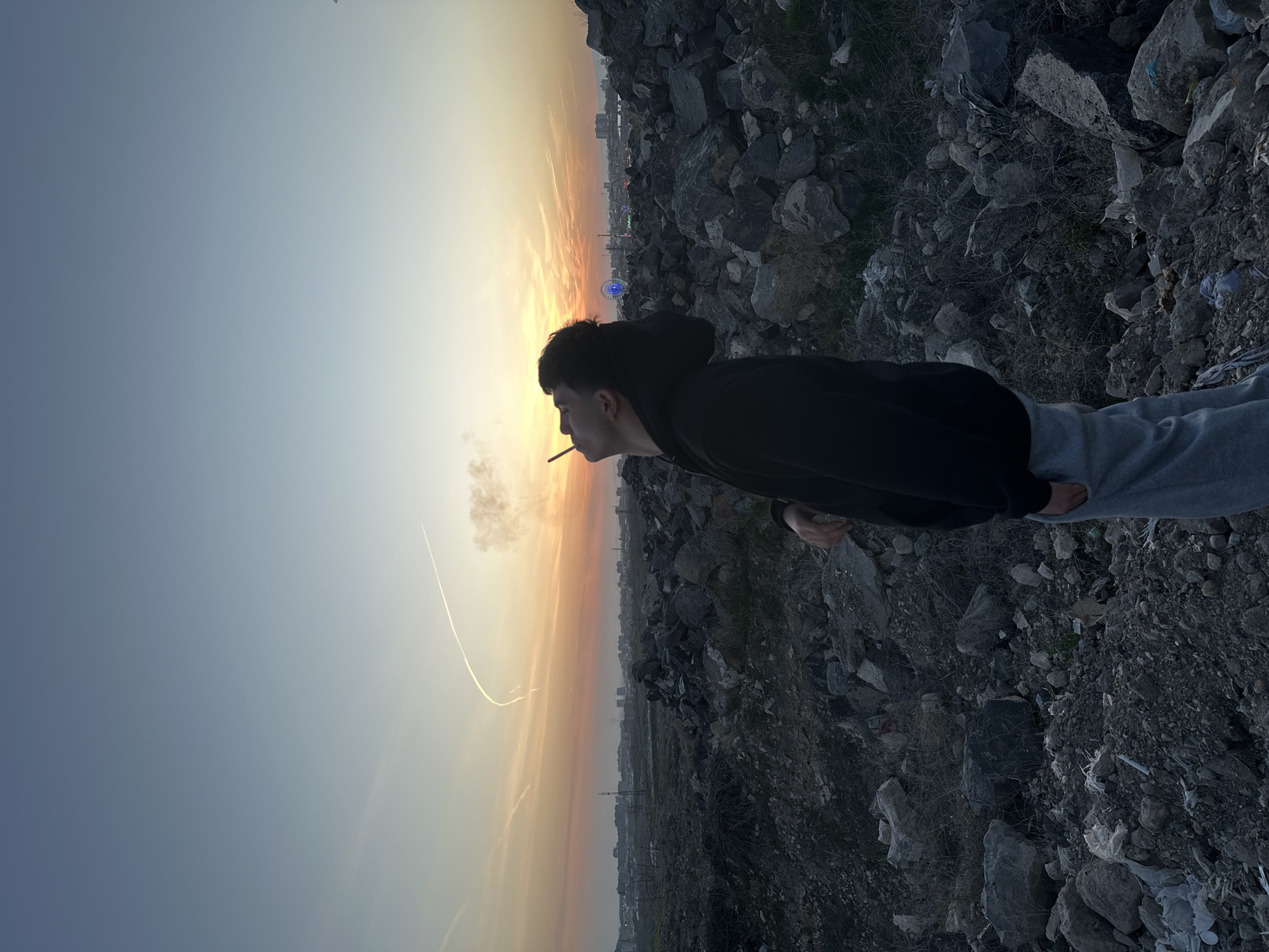 A person is smoking a cigarette against a vibrant sunset backdrop in a rocky area, likely in an urban or suburban setting. The person is wearing a dark hoodie and grey pants, their side profile is visible with the cigarette in their mouth. In the background, there's a city skyline with what appears to be a Ferris wheel illuminated, and the sky transitions from yellow to orange. The ground consists of rocks, dirt, and some sparse vegetation.