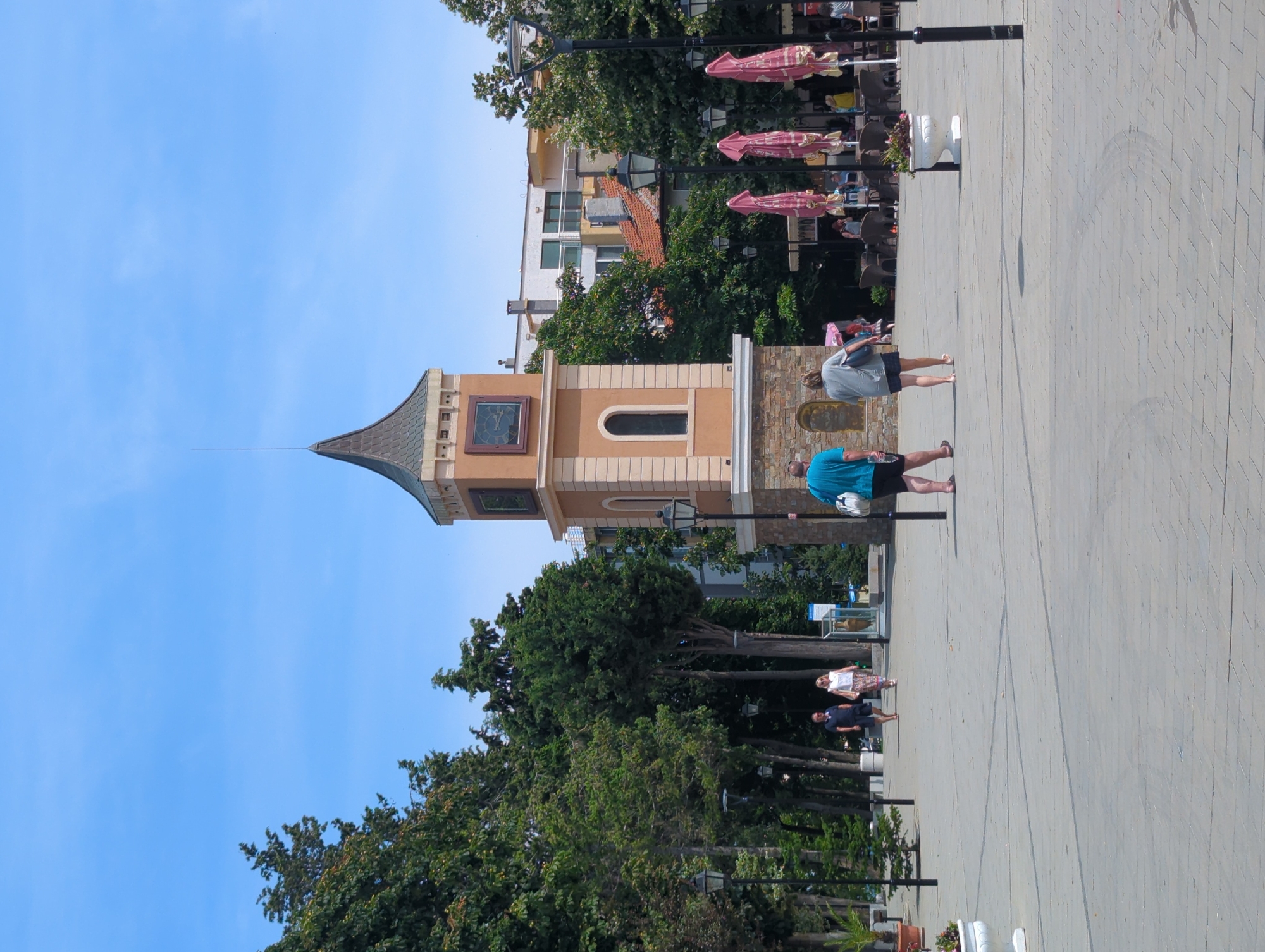This image captures a scene in the city of Obzor, Bulgaria, featuring a prominent clock tower in the center. The tower has a distinctive orange facade with a darker, conical roof and a clock face visible on one side. In front of the tower, a paved plaza is sparsely populated with a few individuals walking. To the left, lush green trees line the area, creating a park-like setting, and several lampposts are strategically placed throughout the plaza. In the background, buildings and more trees are visible, suggesting an urban environment. The sky is a clear, bright blue. To the right, cafe umbrellas are closed, and outdoor seating is arranged, indicating that the establishments are either not in operation at the moment or have recently closed. The overall impression is a tranquil day in a public square.