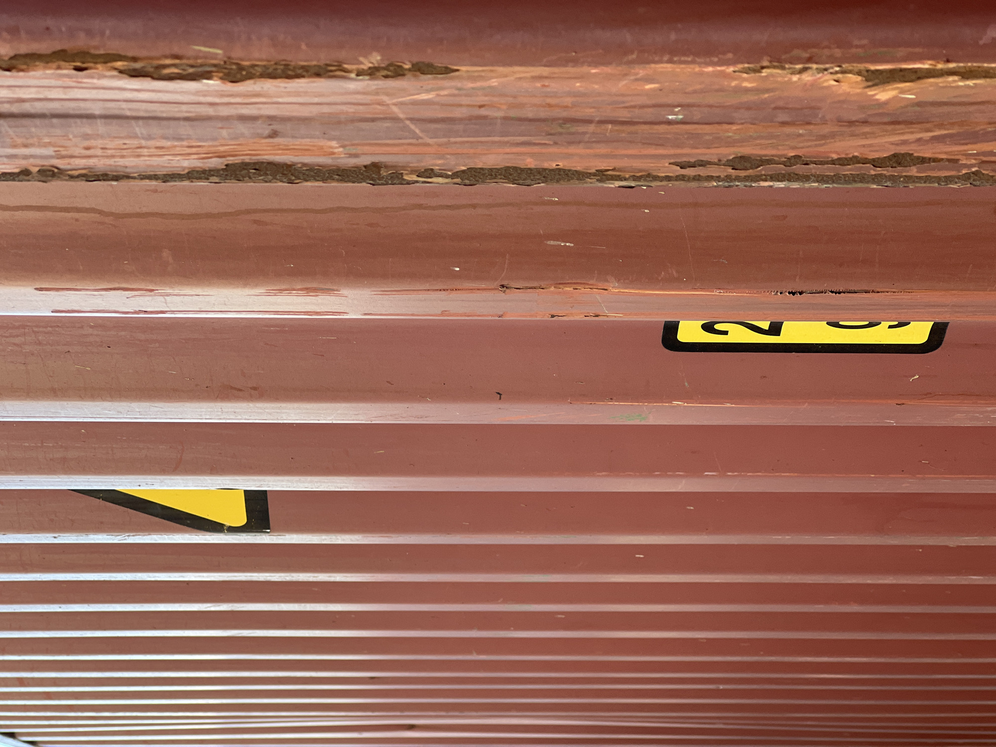 The image shows a close-up, abstract view of the corrugated metal exterior of a shipping container. The perspective is looking upwards, with multiple horizontal ridges of the container dominating the frame. The metal is painted a reddish-brown color, showing signs of wear and tear, including rust patches, scratches, and some dirt.
In the upper part of the image, there are more significant areas of rust and flaking paint, revealing the underlying metal and texture.
Two yellow warning labels with black text and borders are visible. One label, partially visible on the right side, appears to have the numeral "2" and other text. Another yellow triangle with a black border is visible in the lower-middle section of the image.
The overall impression is one of industrial decay and the passage of time. There are no people or clear indications of a specific event. The lighting suggests it is daytime, but no specific time or weather conditions are discernible. The location context provided is Újezd u Brna, Czechia, which, combined with the shipping container, suggests an outdoor industrial or transportation-related setting.