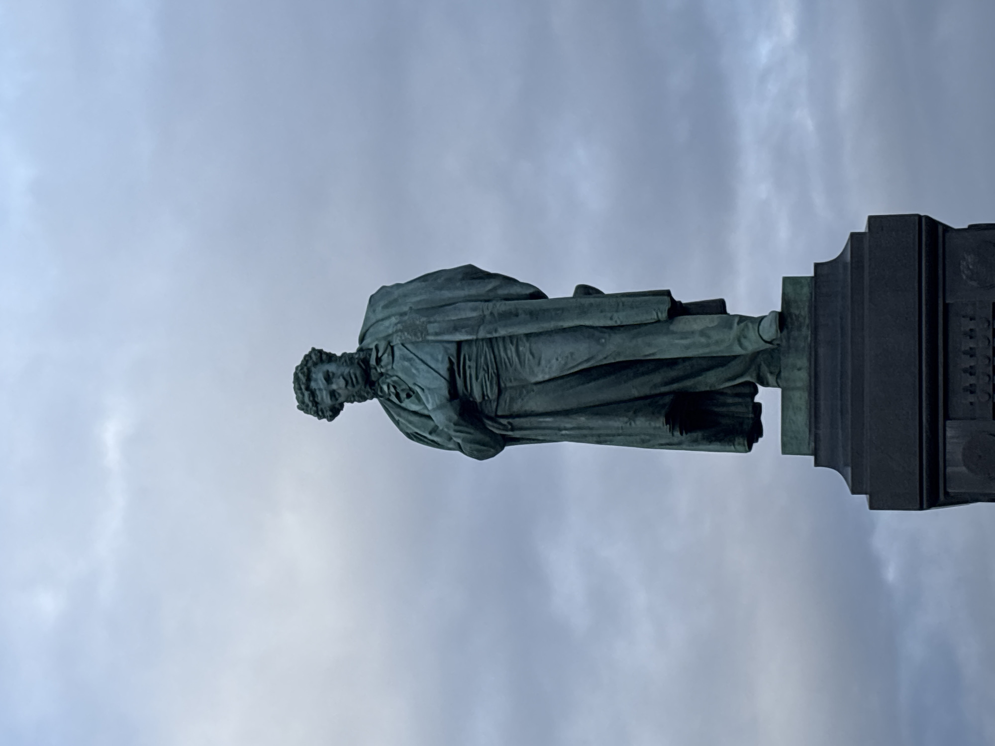 The image shows a bronze statue of Alexander Pushkin in Moscow, Russia. The statue is set against a cloudy sky. Pushkin is depicted in a cloak-like garment. The statue is mounted on a dark-colored, multi-tiered plinth.