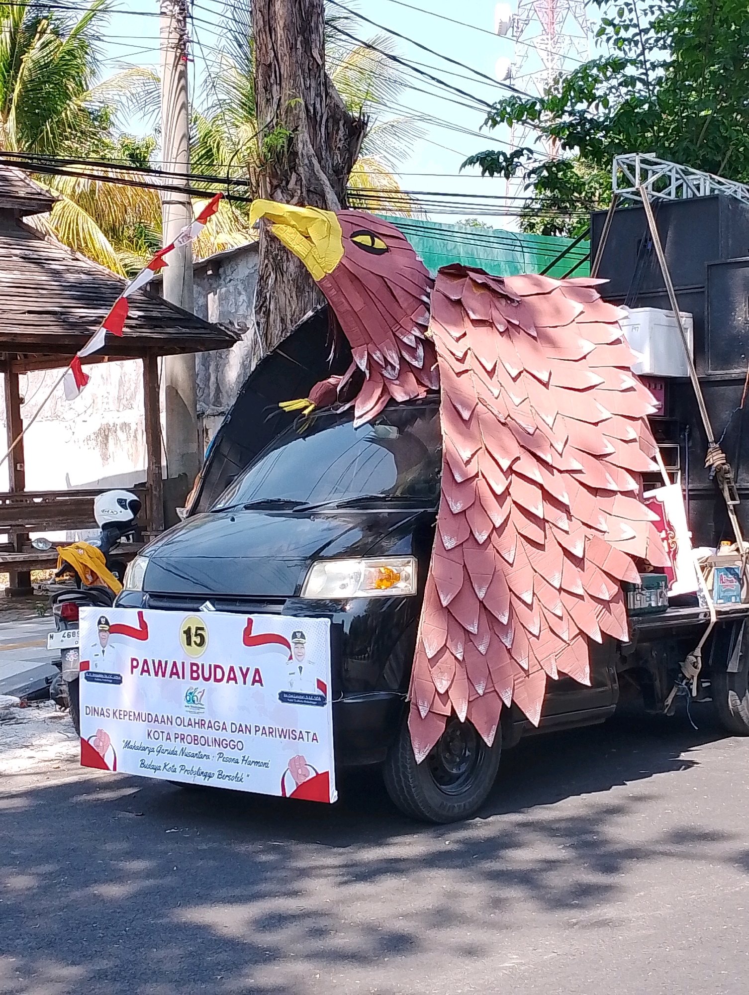 A large, red, eagle-shaped float with yellow accents is mounted on the back of a dark-colored truck. The eagle's wings are spread wide over the truck's cab, and its head is positioned above the windshield. A banner is attached to the front of the truck, displaying text in Indonesian and images of two men in formal attire. The banner reads "PAWAI BUDAYA" which translates to "Cultural Parade", and underneath it states "DINAS KEPEMUDAAN OLAHRAGA DAN PARIWISATA KOTA PROBOLINGGO" (Department of Youth, Sports, and Tourism of Probolinggo City). Below this, a slogan reads, "Maksiya Garuda Nusantara, Pisana Harmoni Budaya Kota Probolinggo Berisol". There is a number "15" in a yellow circle on the banner. The truck is parked on an asphalt road, with trees, power lines, and a distant tower visible in the background. A motorcycle with a white helmet is parked to the left of the truck. The scene appears to be outdoors, possibly during a daytime event or parade in the city of Probolinggo, Indonesia.