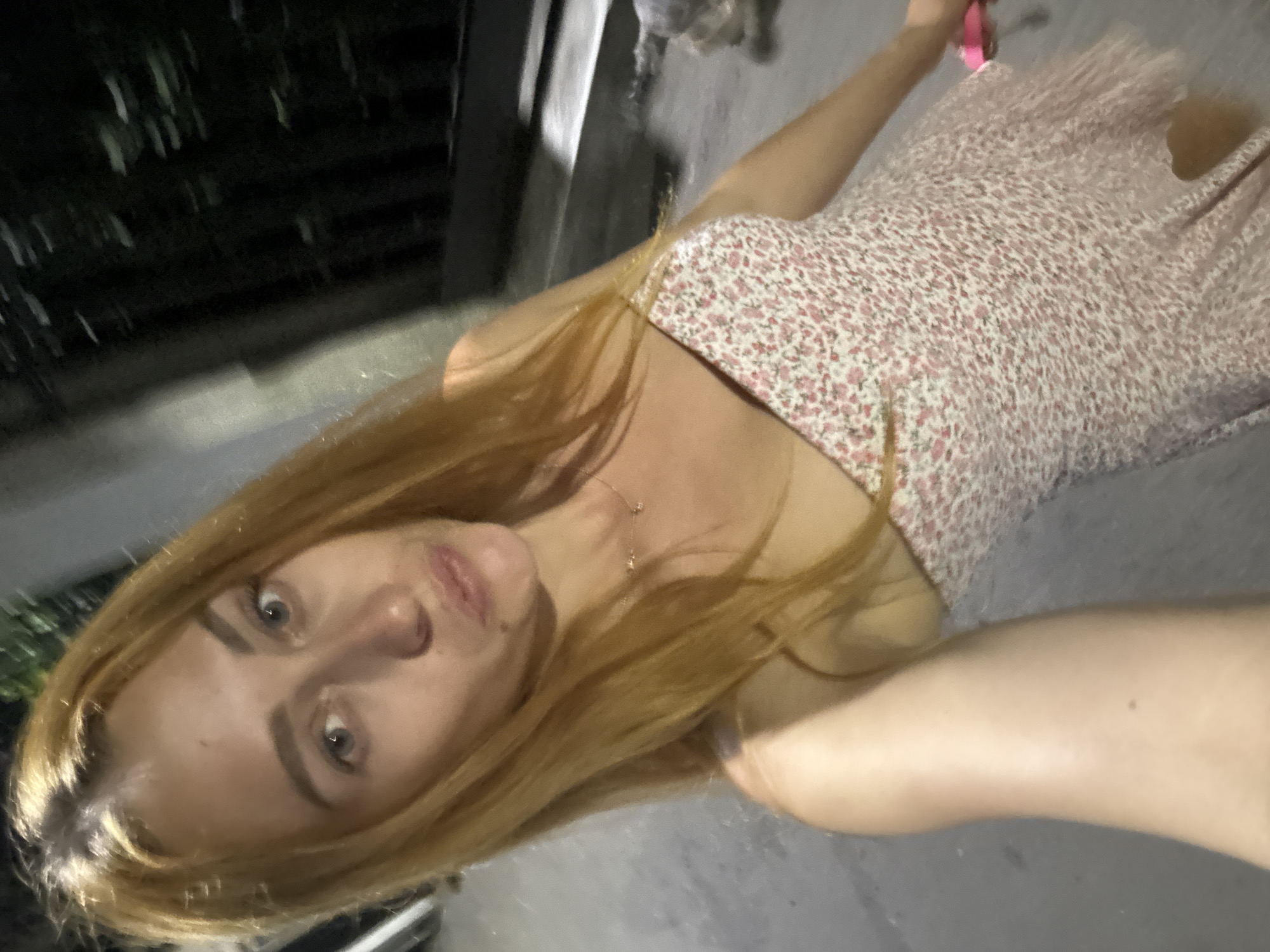 A woman with blonde hair wears a pink floral dress while holding a pink object, possibly a leash. She stands on a paved surface, with blurry trees and structures in the background, suggesting an outdoor urban setting at night. The image was taken in Budva, Montenegro.