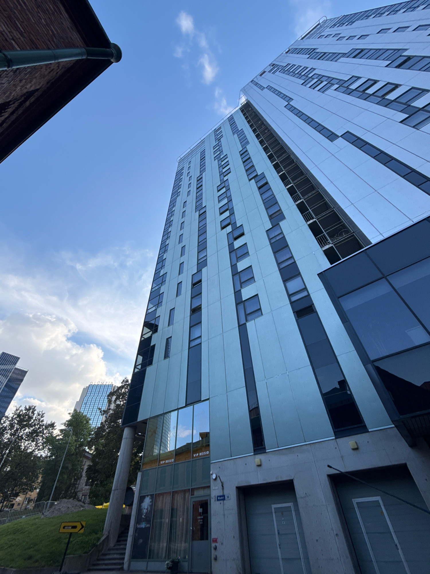 The image shows a low-angle, wide-shot perspective of a modern, tall building in Tallinn, Estonia, against a blue sky with scattered clouds. The building's facade is a light blue-grey color with a regular pattern of windows. The lower part of the building features a darker glass facade and concrete elements, including two large garage doors. Signs on the building indicate "VIP REISID" and "Masku 36". In the background, other modern buildings and lush green trees are visible. The overall impression is one of contemporary urban architecture.