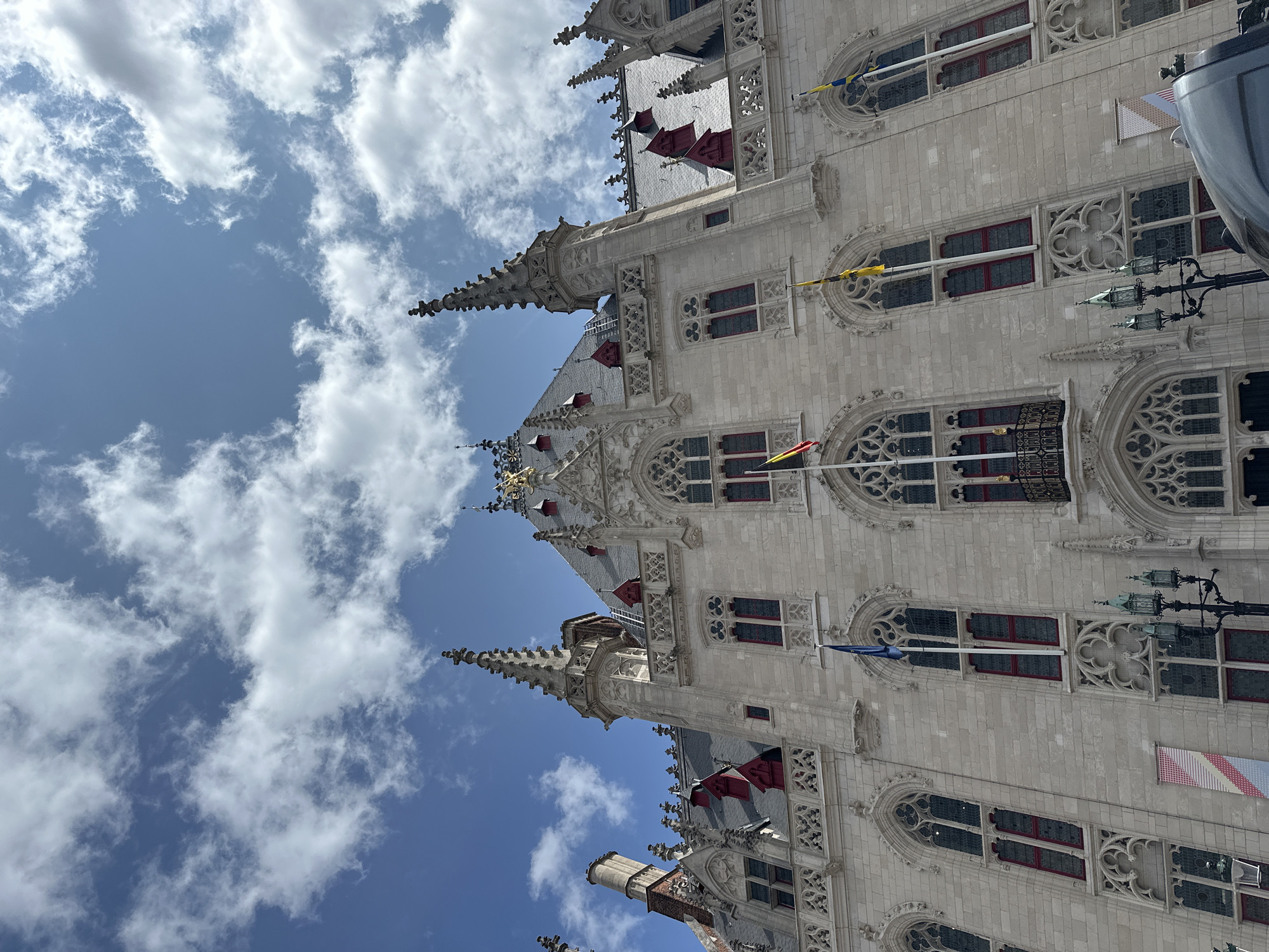 The image shows the facade of a grand building, likely a historic government building or landmark, in Bruges, Belgium. The architecture is Gothic, characterized by pointed arches, intricate stonework, and multiple spires. The building is constructed of light-colored stone and features numerous tall, narrow windows with decorative tracery. Some windows have red frames. Several flags are visible, including what appear to be the flags of Belgium and possibly Flanders. Red dormer windows punctuate the grey slate roof. The sky above is a bright blue with scattered white clouds. The perspective is looking up at the building from a low angle, emphasizing its height and grandeur. The lower portion of the image shows a glimpse of a dark-colored vehicle and a lamppost with multiple lanterns, suggesting the building is situated in a public square or street. There are no people clearly visible in the frame, but the presence of the vehicle and lamppost indicates it is a public space that is in use. The overall impression is one of historical significance and architectural beauty.
