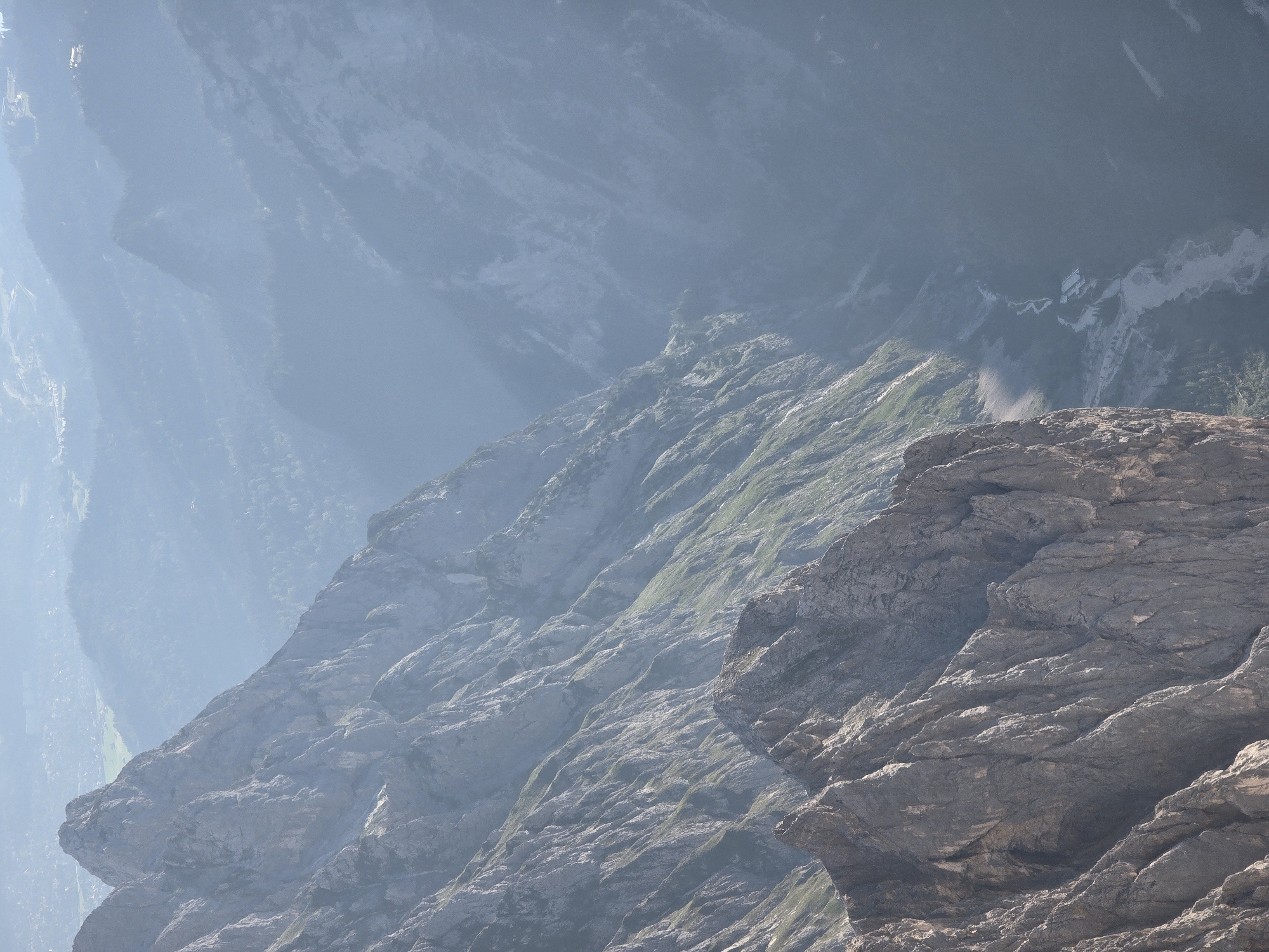 The image depicts a vast, mountainous landscape, likely in the vicinity of Grainau, Germany, given the distinctive rock formations and the presence of a valley with a river. In the foreground, a jagged, imposing rock face dominates the left side of the frame, showcasing intricate textures and steep, weathered slopes. Further back, a series of imposing mountain ridges stretch into the distance, their upper sections shrouded in a light, atmospheric haze. The slopes are a mix of exposed rock and sparse vegetation, with patches of green clinging to the inclines.
A deep valley is visible to the right of the mountains, carved by a winding river that appears to be a mix of water and gravel. Several small structures, possibly buildings or a small hamlet, are situated near the riverbed, nestled within the valley. The background reveals a layering of mountain ranges, increasingly softened by the atmospheric perspective, with hints of a distant settlement or town visible on the far left. The overall impression is one of rugged, natural grandeur, with the harshness of the rock softened by the ethereal quality of the mist. There are no people or animals visible in the image.
