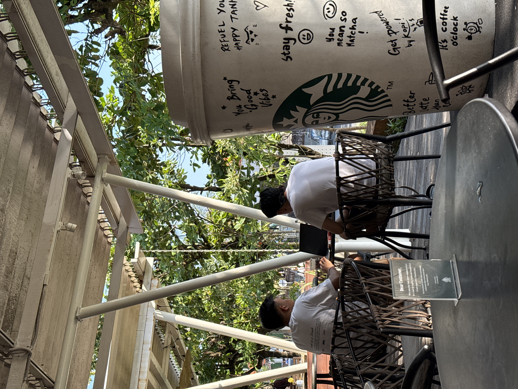 An outdoor daytime scene depicts a vibrant cafe setting, likely in Kuta, Indonesia, with two men seated at separate tables. The environment is bright and sunny, featuring white architectural beams and an awning, with a clear blue sky peeking through lush green tree foliage. Both men are dressed in white T-shirts and appear to be engaged with laptops or devices.
A dominant feature in the foreground is a large, oversized replica of a Starbucks coffee cup, which is covered with various hand-drawn inspirational phrases and doodles in black marker. Clearly visible text on the cup includes: "REFUEL YOU HAPPY TANK", "Stay fresh!", "You Bring Good Vibes", "You mean so Matcha !", "Get powered by Kopi!", "It's coffee o'clock", and a partial "Better latte than never". The iconic Starbucks siren logo is also prominently displayed on the cup.
The men are seated on dark wicker-style chairs at dark-surfaced tables. On one table, a small white sign is present, partially readable as "Dear Customer,". The atmosphere suggests a relaxed, casual outdoor workspace or leisure spot, with the tropical greenery adding to the ambiance.
