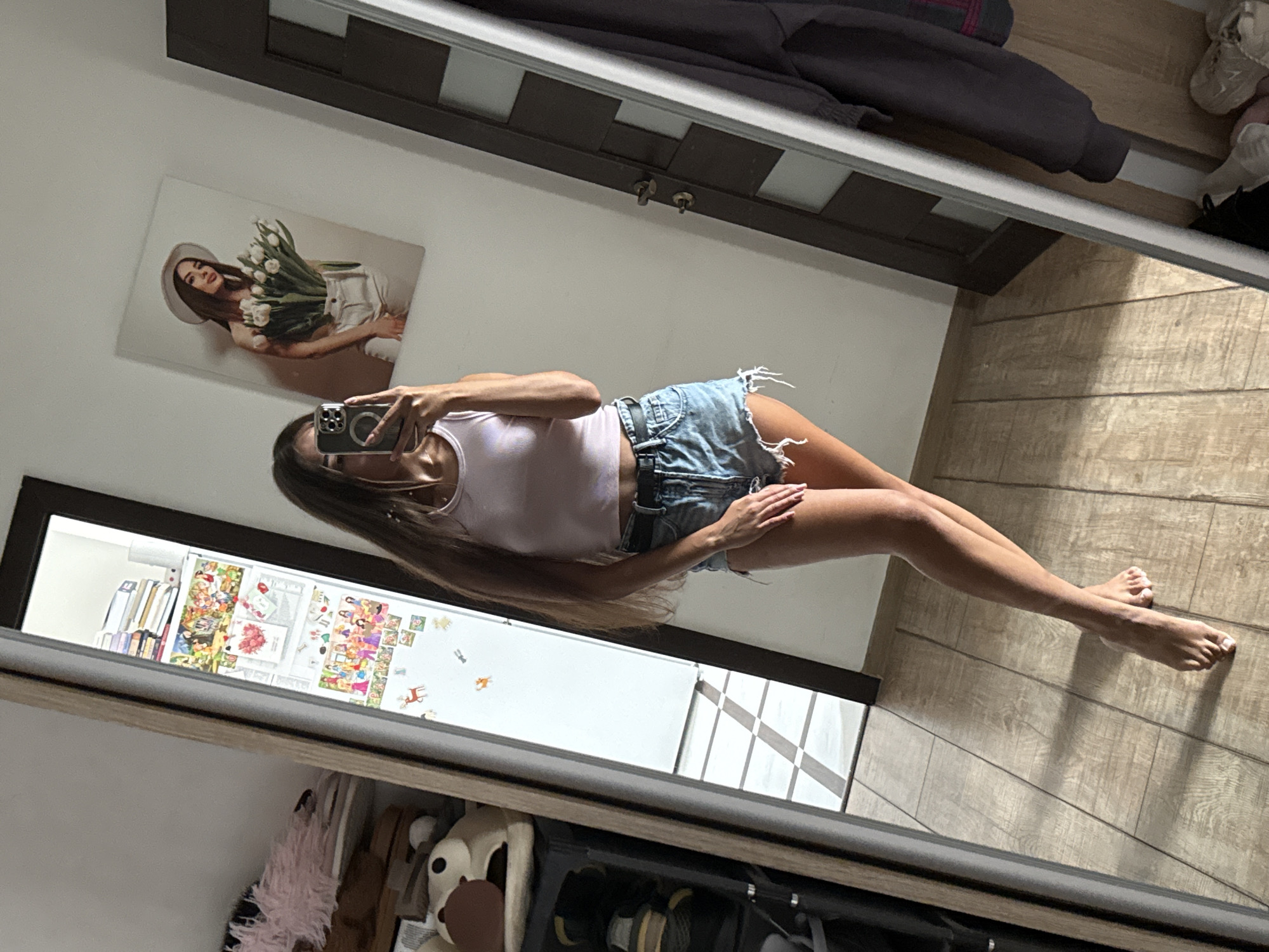 A full-body mirror selfie is taken by a young woman with long, dark brown hair, wearing a light pink crop top and distressed denim shorts. She is standing in a room with light wooden flooring and a white wall. To the left of the frame, a tall mirror reflects a portion of the room, including shelves with books, toys, and a dark shoe rack. On the wall, a framed picture of a woman holding tulips is visible. To the right, a dark door with a geometric pattern is present, and a dark grey garment hangs on a hook. The woman's pose is casual, with one leg slightly bent and crossed in front of the other, and her feet are bare. The lighting suggests it is daytime, with shadows cast on the floor. This scene is likely a residential interior in Chișinău, Moldova, given the presence of a framed artwork and personal items typically found in a home environment.