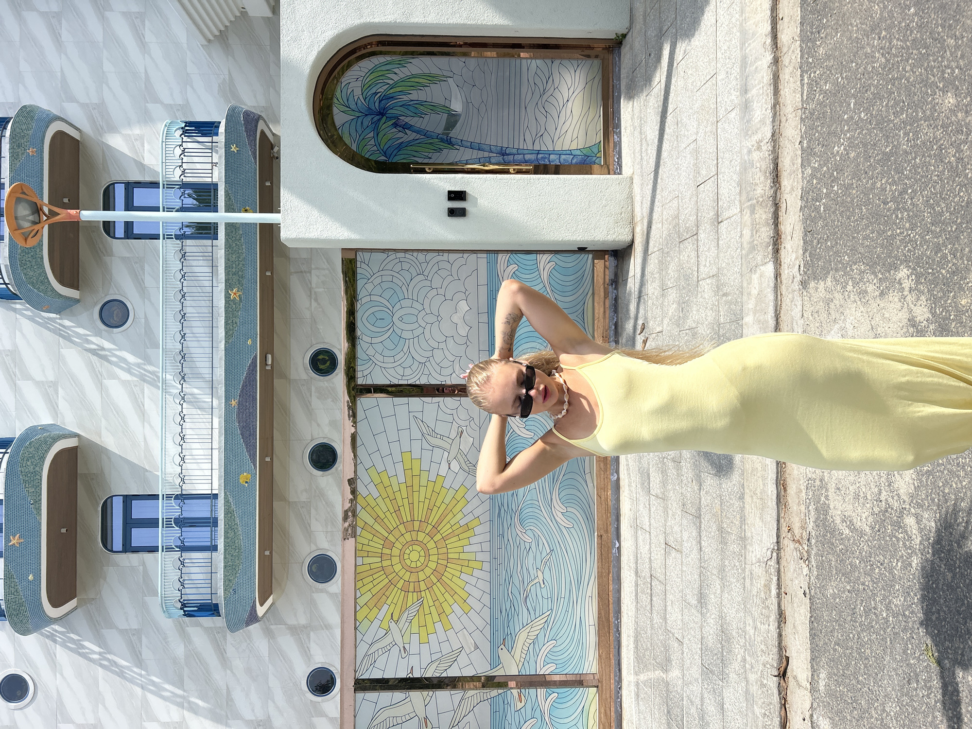A young woman with blonde hair pulled back and wearing sunglasses, a light yellow tank dress, and a pearl necklace stands with her hands behind her head in front of a building. The building has a white, marble-like facade with balconies adorned with blue railings and mosaic tile details featuring marine life. Below the balconies, a large stained-glass mural depicts a bright yellow sun with rays, seagulls in flight, and a stylized ocean with waves. To the right of the mural is an arched entryway with a stained-glass panel featuring a palm tree. The ground in front of the building is paved with grey stone tiles and asphalt. The scene is set on a sunny day, with shadows cast on the ground. This location is in Nam Nha Trang Ward, Vietnam.