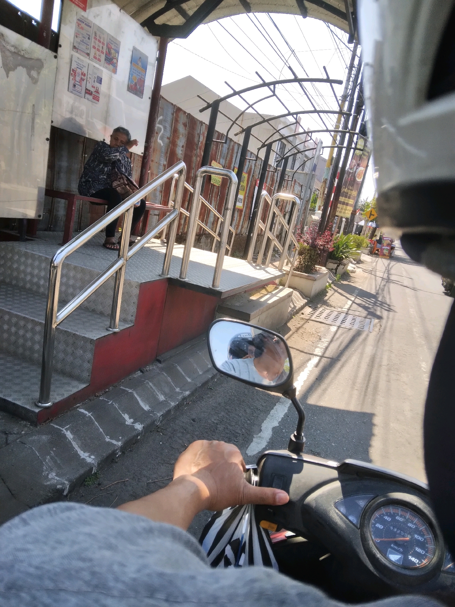 The image is taken from the perspective of someone riding a motorcycle in Purwokerto, Indonesia. In the foreground, the hand of the rider is visible gripping the handlebar, with a partial view of the motorcycle's dashboard which shows the speedometer reading approximately 40 km/h. The rider is wearing a gray long-sleeved shirt. A side mirror reflects the rider's helmeted head.
In the mid-ground, a covered bus stop or waiting area is situated alongside a street. A woman with dark hair and a patterned blue and white shirt is sitting on a red bench within the bus stop, her head resting on her arm as if sleeping or resting. The bus stop features a metal roof supported by posts, corrugated metal walls, and a staircase with stainless steel railings leading up to a platform. The steps are covered in a textured metal material.
The background shows a street with other buildings and signs, indicating an urban environment. Overhead, a complex network of electrical wires is visible against the bright sky. The sunlight casts strong shadows on the road, suggesting it is a sunny day. The overall scene depicts a typical street view in an Indonesian city, likely during daylight hours.