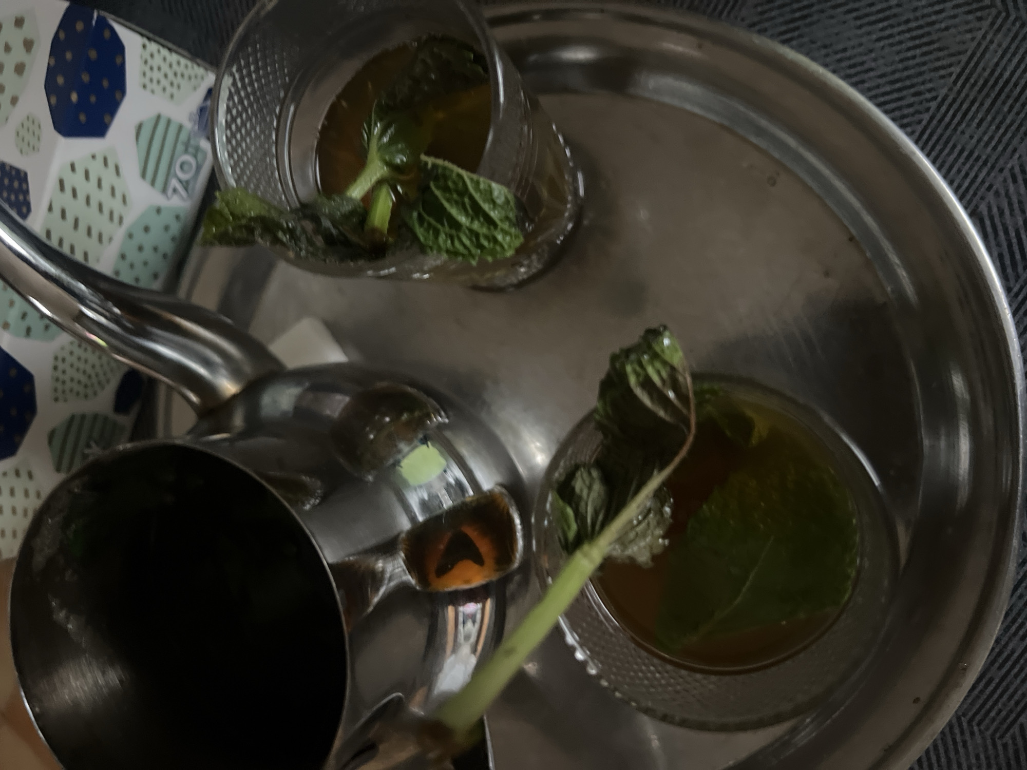 This image, captured in Casablanca, Morocco, presents a traditional Moroccan mint tea service setup. A reflective, silver-colored metal tray occupies the central foreground, holding a polished metal teapot and two clear glass tea cups. The teapot's interior appears dark, consistent with containing liquid. Each glass contains a transparent, amber-brown liquid, identified as tea, with several sprigs of fresh green mint leaves submerged within. One glass exhibits a textured pattern on its exterior, and a long green stem, possibly an additional herb or a larger mint stalk, extends from it towards the teapot. The tray's surface shows minor smudges and reflections. In the blurred background, a patterned object, likely a tissue box, displays a geometric design of blue, teal, and green polygons against a white background; the number "70" is faintly visible on one teal section. The tray rests on a dark, textile surface featuring a subtle diagonal striped pattern. No individuals are present in the scene.