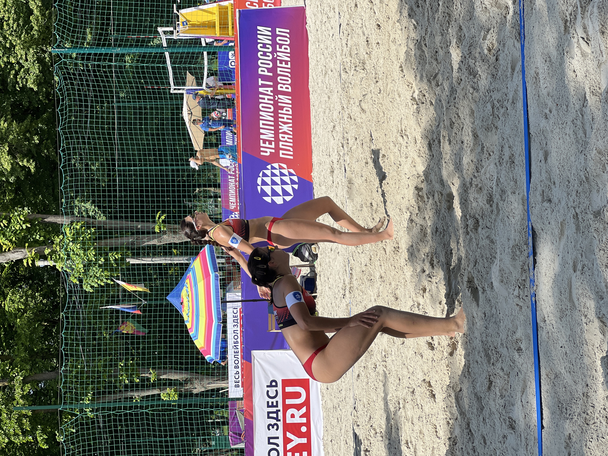 The image shows two women playing beach volleyball. They are both wearing red and dark red bikinis, and they are positioned on the sand, ready to receive the ball. In the background is a banner in Russian advertising a volleyball tournament. A net is also visible behind the banners, along with an umbrella and flags. The beach volleyball court is located outdoors, with trees visible in the background. Based on the text on the banner, the image is from Russia, specifically Krasnodar.