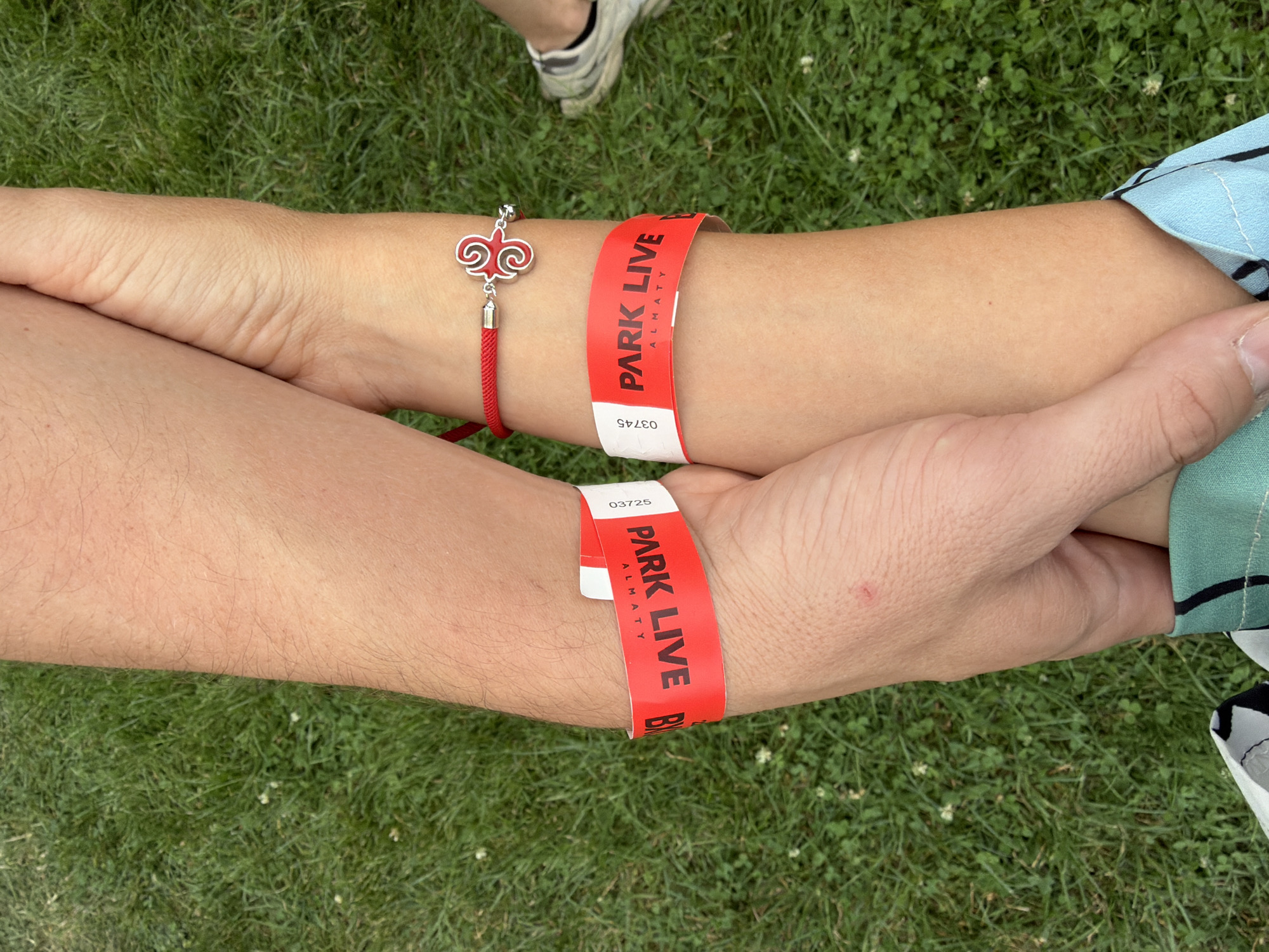 The image shows a close-up, overhead view of two people holding forearms outdoors on a grassy area. Both individuals are wearing red wristbands with black text that reads "PARK LIVE" and "ALMATY", along with numbers. The wristband on the left arm reads "PARK LIVE B" and has the number "03725" visible. The wristband on the right arm reads "PARK LIVE ALMATY" and has the number "03745". The person on the right also wears a red string bracelet with a silver charm resembling a stylized heart or key. The grassy environment suggests an outdoor event or park setting. The image does not provide enough information to determine the specific location within Pervomayskiye Prudy, Kazakhstan.