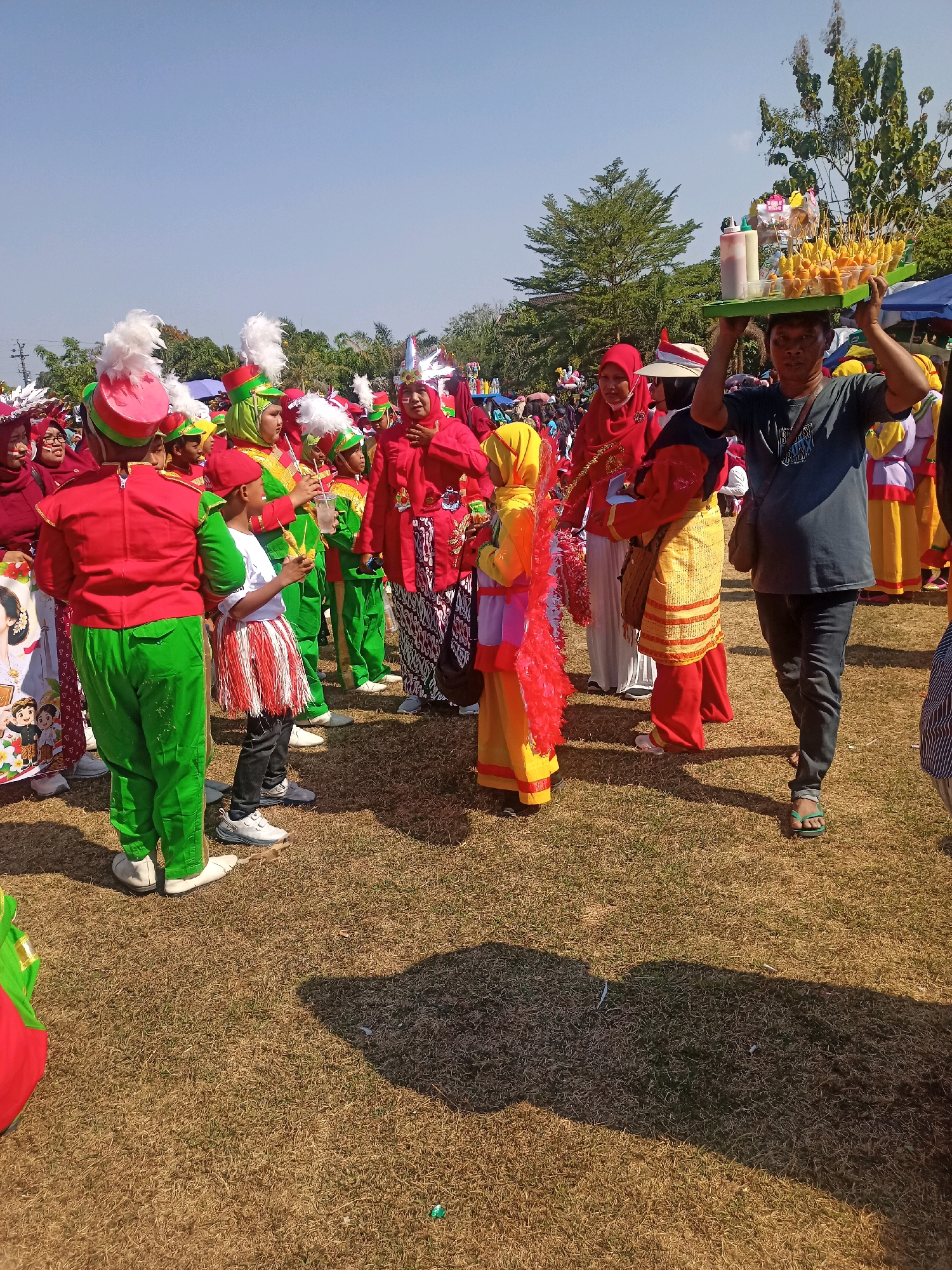 This image captures a vibrant outdoor gathering, likely a parade or festival, in the city of Sumberlawang, Indonesia. The scene is dominated by a group of people, many of whom are dressed in colorful marching band uniforms. These uniforms consist of red jackets with gold accents, green pants, and festive hats adorned with white feathers. Some individuals are wearing red headscarves.
In the foreground, a young boy in a white t-shirt and a skirt with red and white stripes stands holding a small cup with yellow items on sticks. To his right, a man is carrying a tray of food items, possibly snacks, on his head, balanced by his hands. He is wearing a dark t-shirt and dark pants, and green flip-flops.
A variety of other people are visible in the background, including women in red headscarves and other individuals in similarly festive attire. The ground is covered in dry, brown grass, suggesting an outdoor location, possibly a field or park. The sky is clear and blue, indicating a sunny day. A few trees are visible in the distance, as are parts of what appear to be tents or stalls. The overall atmosphere seems lively and celebratory.