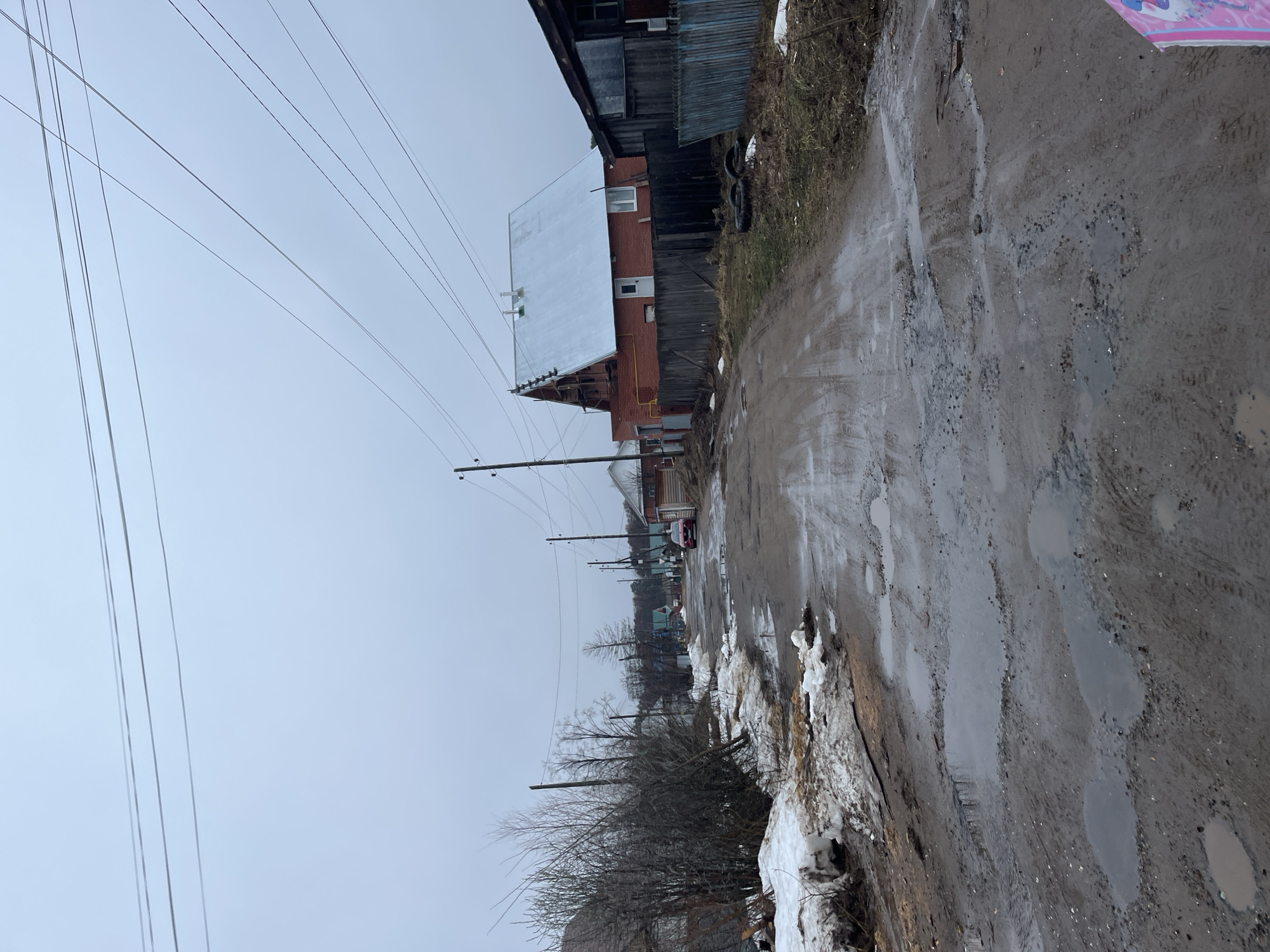 The image depicts a muddy, potholed road in what appears to be a rural or semi-rural area. The road is wet and contains numerous puddles, with patches of melting snow visible along the edges. A building constructed of brick with a metal roof, and adjacent wooden structure is seen on the right side of the road. Power lines stretch across the sky above. A distant red car is visible further down the road. The sky is overcast, suggesting a cold and wet day. The location is presumably Chaikovsky, Russia, based on the prompt's specification. No people are visible in the image.